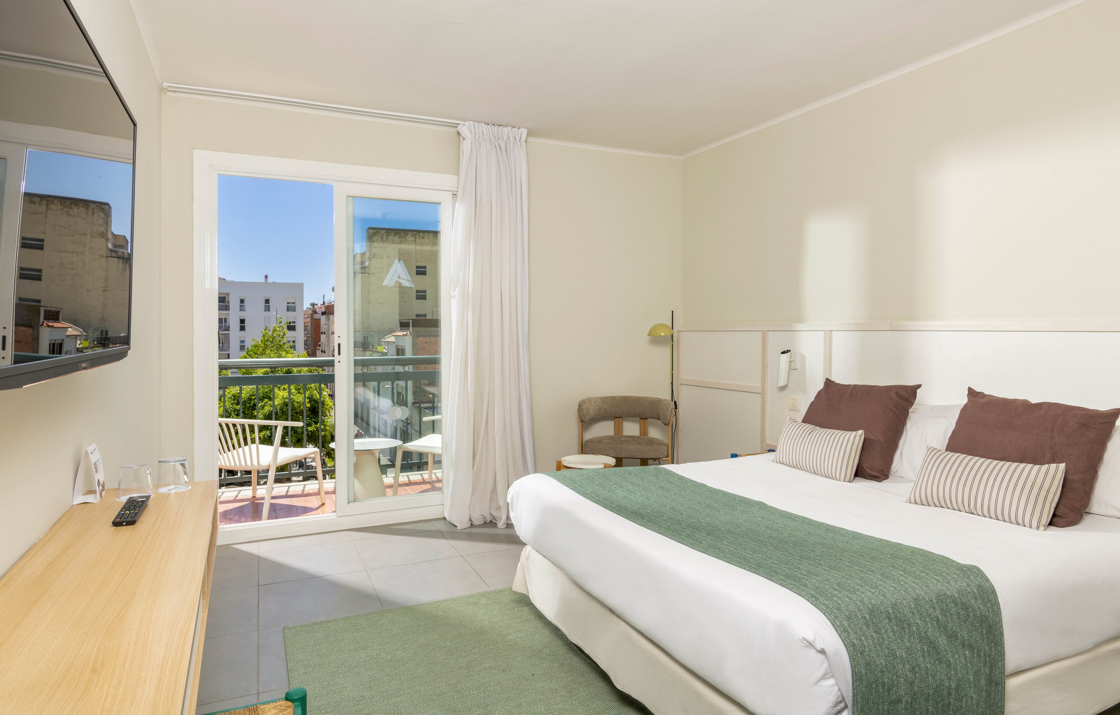 A bright, modern hotel room features a neatly made double bed with white linens and green accents. Large glass doors open onto a small balcony with outdoor chairs, overlooking nearby buildings and greenery. A flat-screen television is mounted on the wall opposite a light wooden desk, and a simple armchair sits in the corner. Soft neutral tones and natural light create a calm, inviting atmosphere.