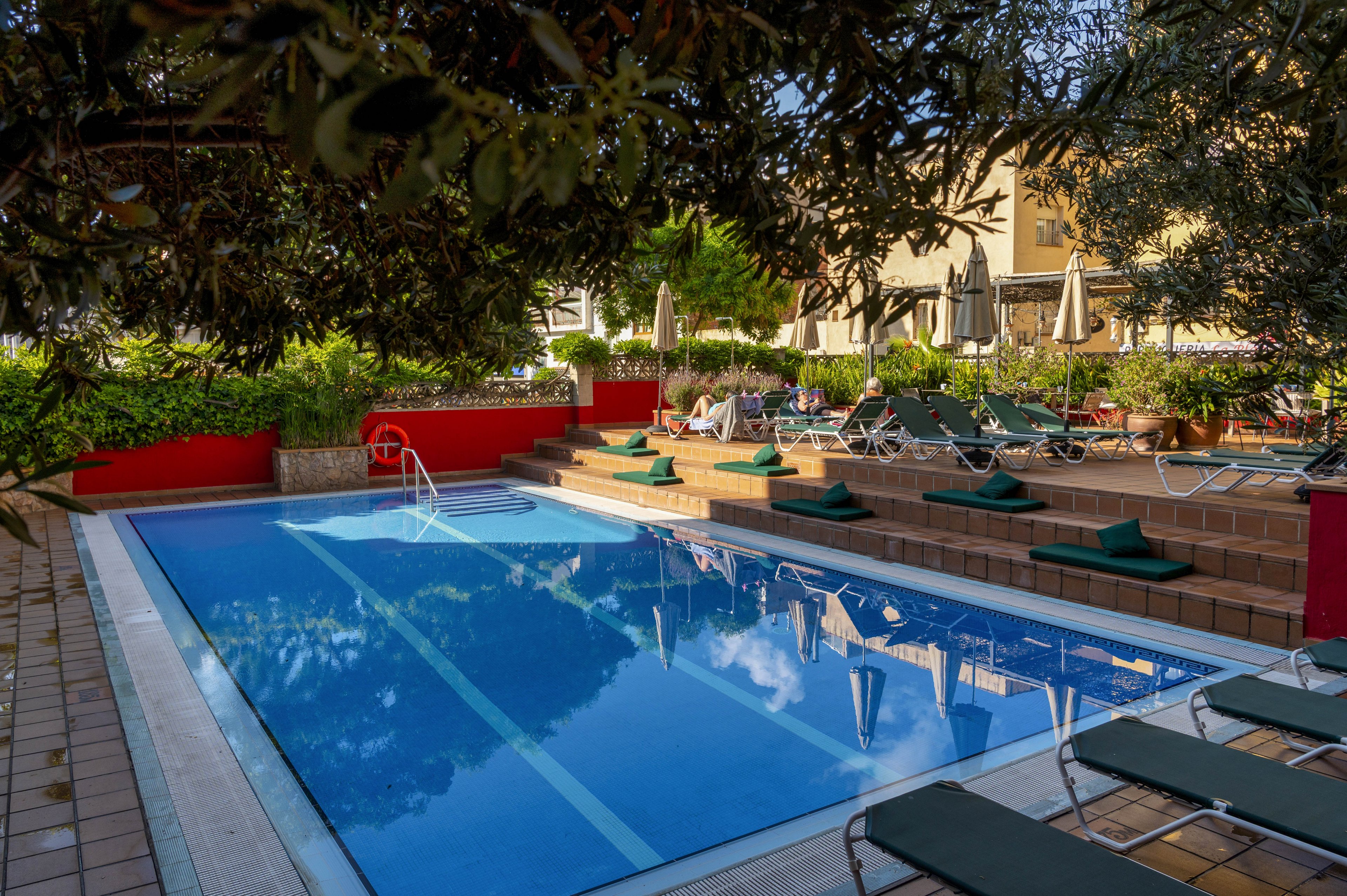 A clear blue swimming pool is surrounded by neatly arranged sun loungers and green cushions in an outdoor hotel courtyard. The pool area is framed by tiled decking, red walls, and lush greenery, creating a secluded retreat. Umbrellas and additional seating are visible in the background, suggesting a comfortable space for guests to relax. Soft natural light and reflections on the water contribute to a calm, inviting atmosphere.