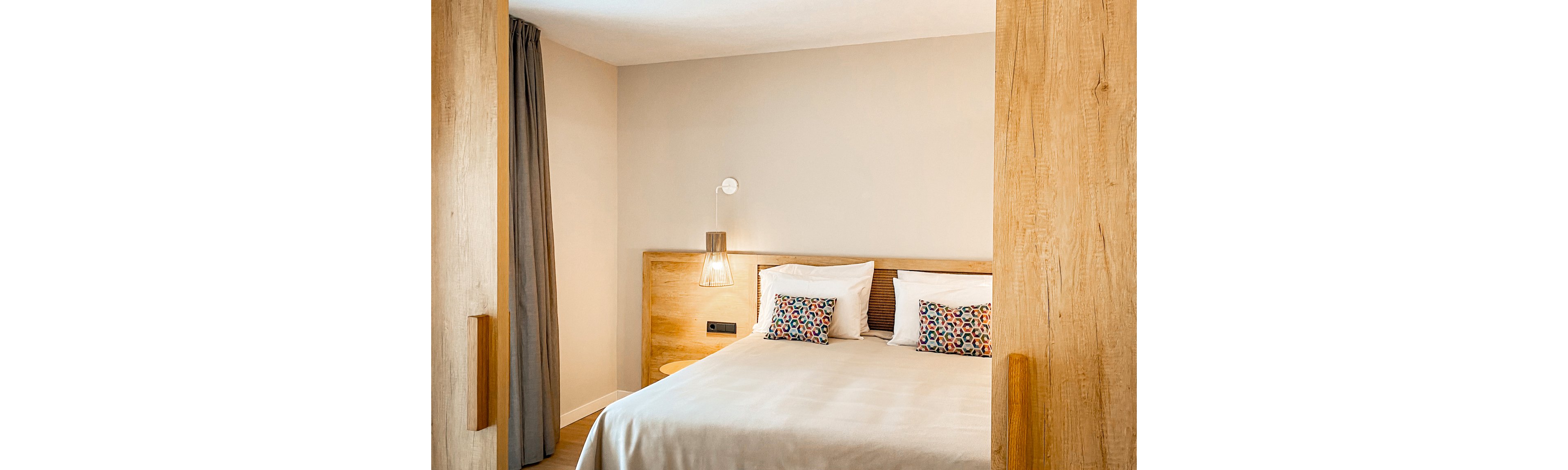 A contemporary hotel room featuring a neatly made double bed with white linens and colorful patterned pillows. The setting includes wooden accents on the bed frame and sliding doors, complemented by soft lighting and neutral walls. A single bedside lamp adds warmth to the minimalist design.