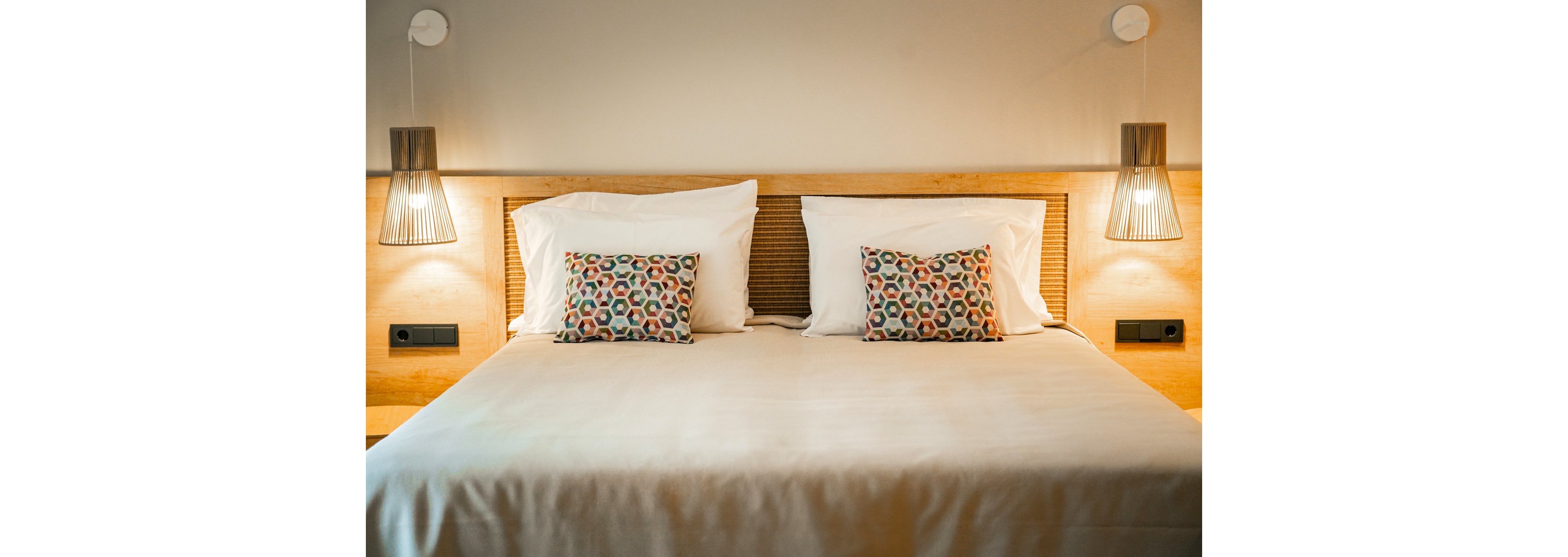 A neatly arranged double bed with white linens and colorful patterned pillows in a modern bedroom. The setting features warm wooden accents and soft ambient lighting from two hanging lamps. The room exudes a calm and inviting atmosphere, ideal for relaxation.