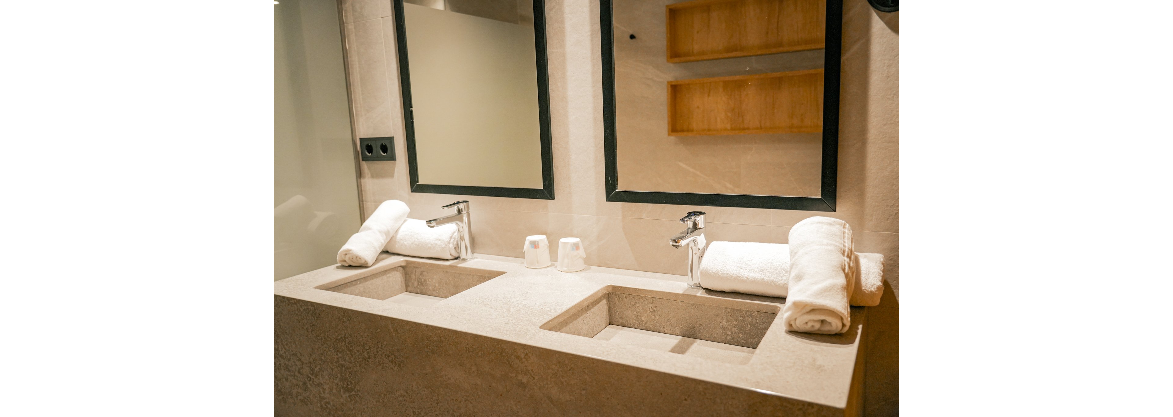 A contemporary bathroom featuring dual rectangular sinks with chrome faucets and matching mirrors. The setting includes neatly rolled white towels and wooden shelves for storage. The neutral beige palette creates a clean and minimalist aesthetic.