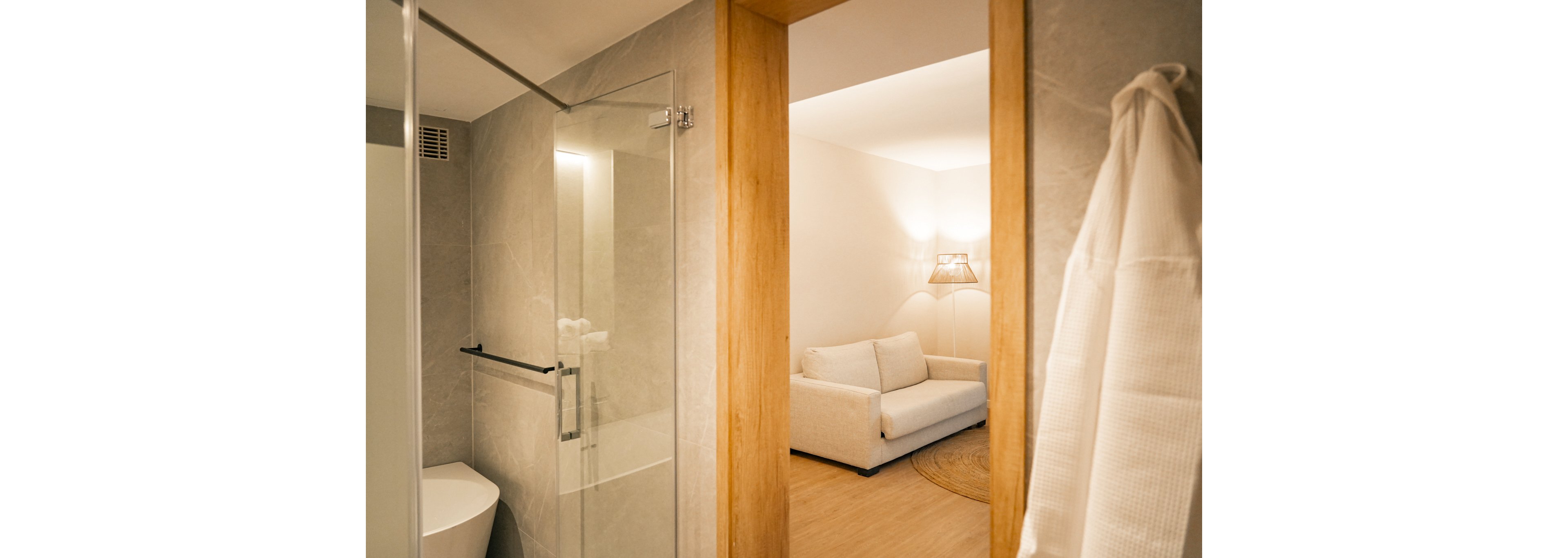 A contemporary bathroom with a glass shower door and sleek fixtures opens into a warm living area. The living space features a white sofa, wooden accents, and soft lighting from a woven lamp. Neutral tones dominate the palette, creating a calm and inviting atmosphere.