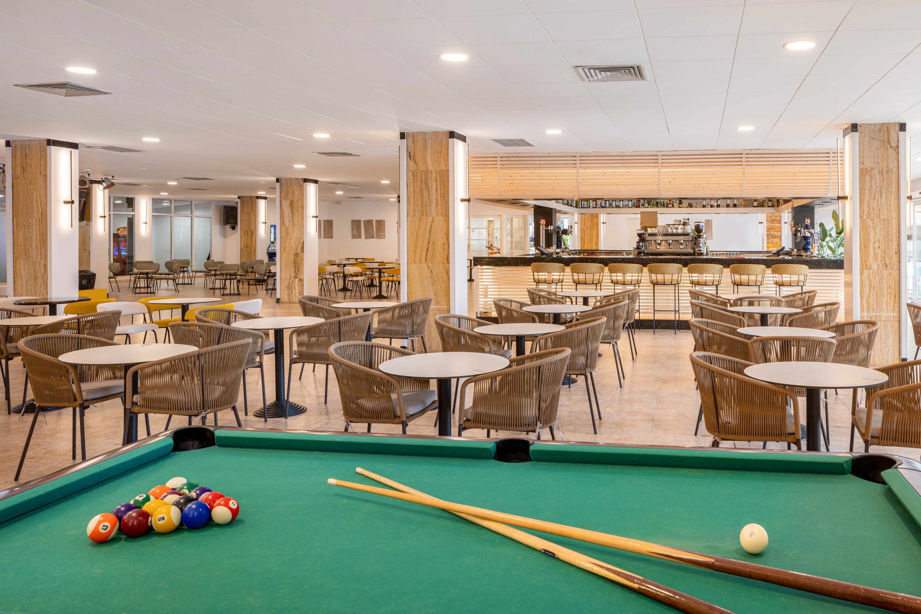 The main subject is a green billiards table with pool balls and cues in the foreground. The setting is a spacious cafeteria or lounge area featuring numerous round tables and wicker chairs, with a bar counter visible in the background. The space is brightly lit with neutral tones and a contemporary style, creating a welcoming and relaxed atmosphere. No visible text or numeric values are present in the image.