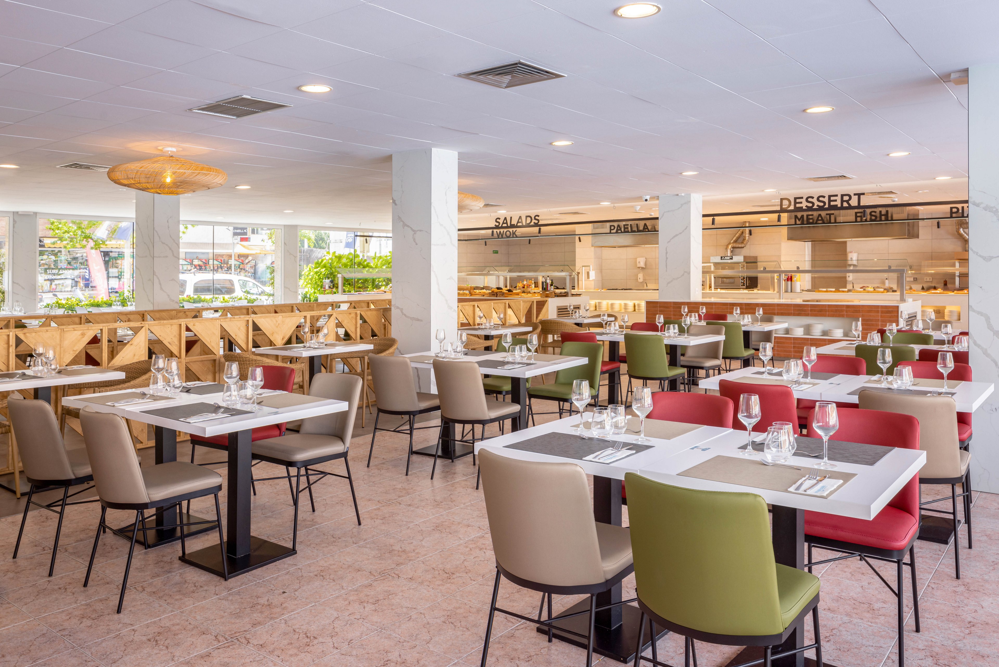 A spacious, contemporary restaurant interior features neatly arranged tables set with glassware and cutlery. The seating includes a mix of olive green, beige, and red chairs, creating a vibrant yet inviting atmosphere. The open kitchen area in the background displays clear signage for salads, desserts, and main dishes. Natural light streams in through large windows, enhancing the clean and modern aesthetic.