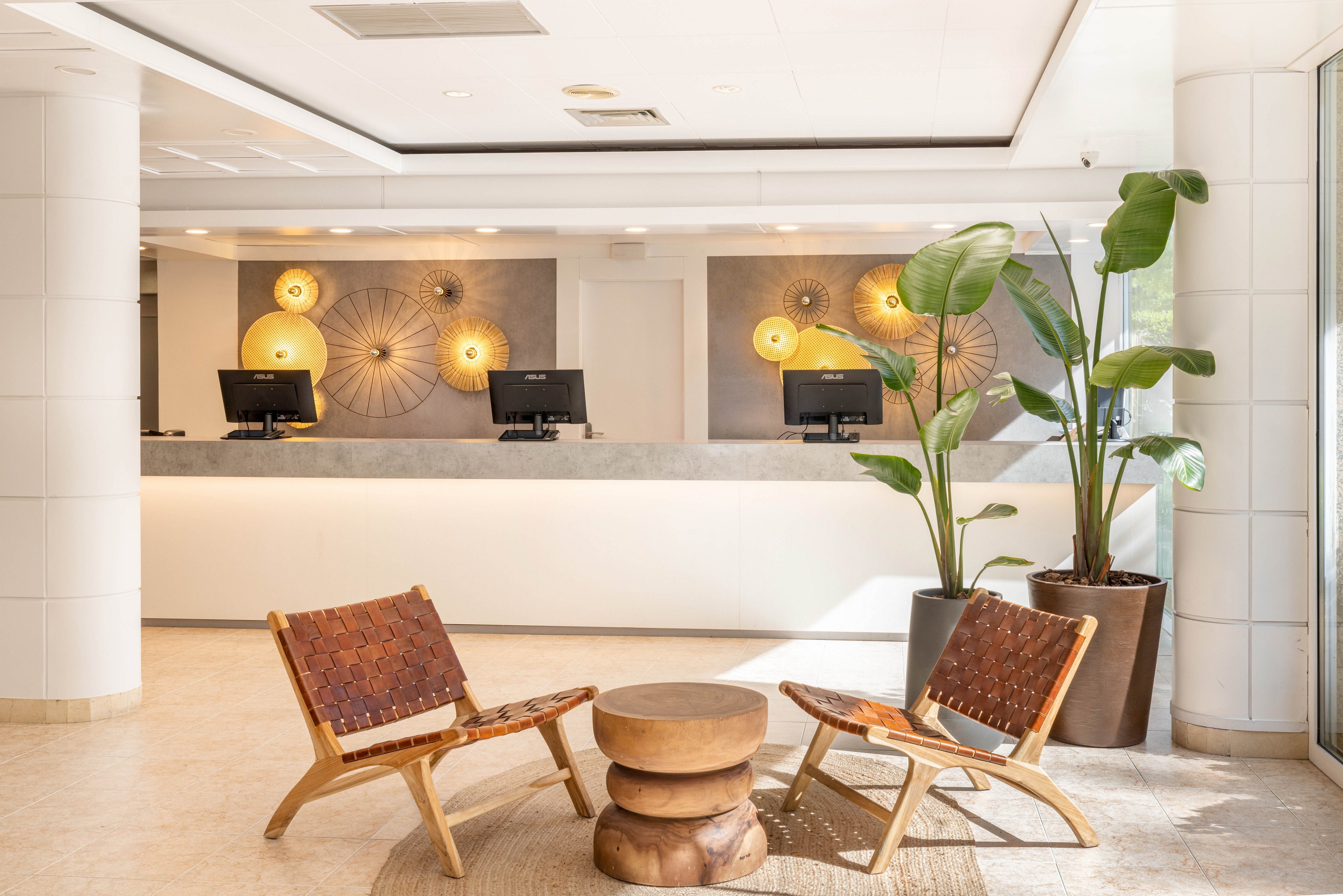 A contemporary hotel lobby features two receptionists standing behind a sleek counter with three computer monitors. The setting is bright and welcoming, with decorative wall lights and large potted plants adding to the ambiance. Two woven chairs and a wooden table are arranged in the foreground, creating a comfortable waiting area. The overall mood is professional and inviting, with neutral tones and natural textures.