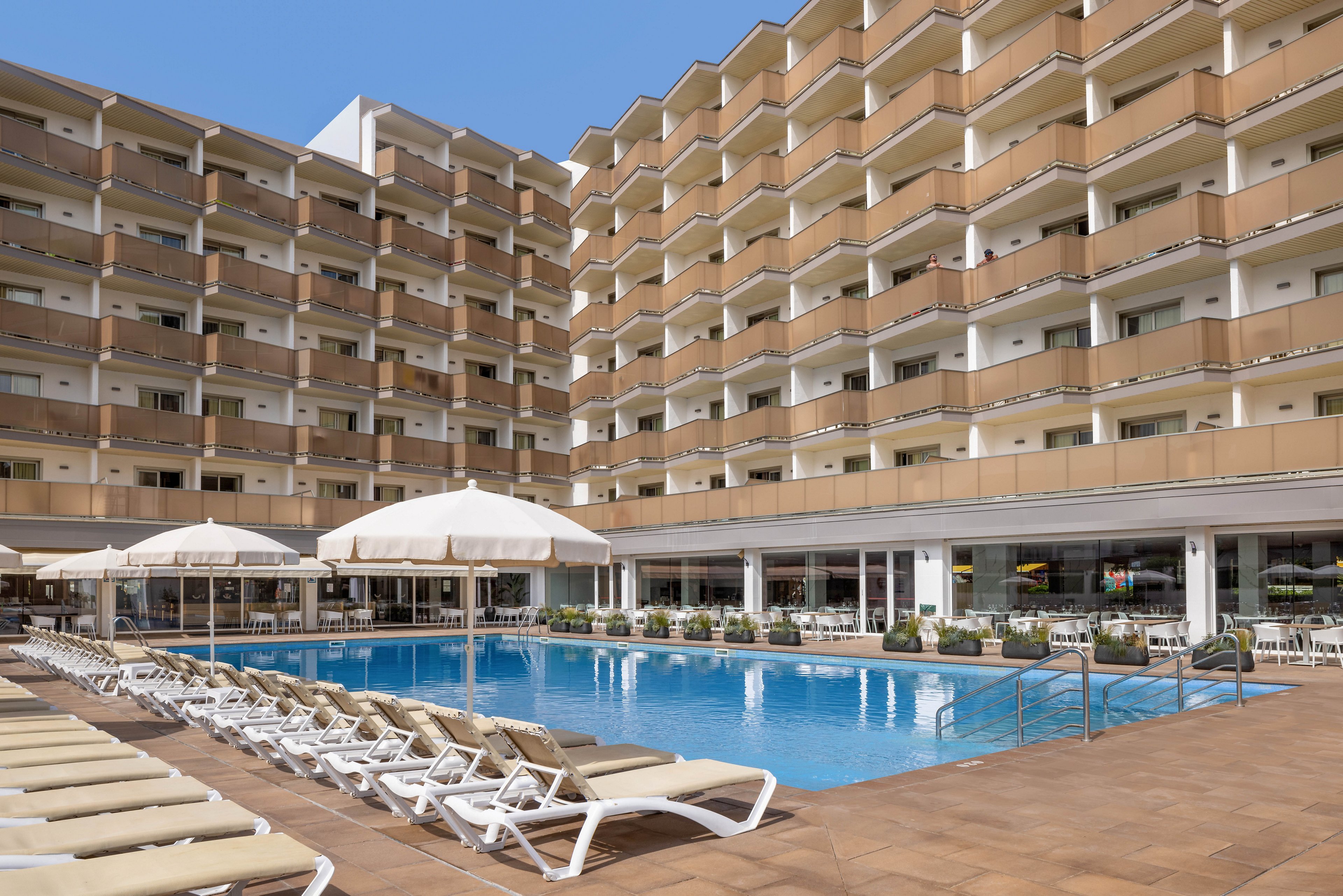 The image shows a contemporary multi-story hotel building with balconies overlooking a large outdoor swimming pool. Numerous white sun loungers are arranged neatly around the pool, and several large umbrellas provide shade. The setting is bright and sunny, with clear blue skies, creating a relaxed and inviting atmosphere for guests.