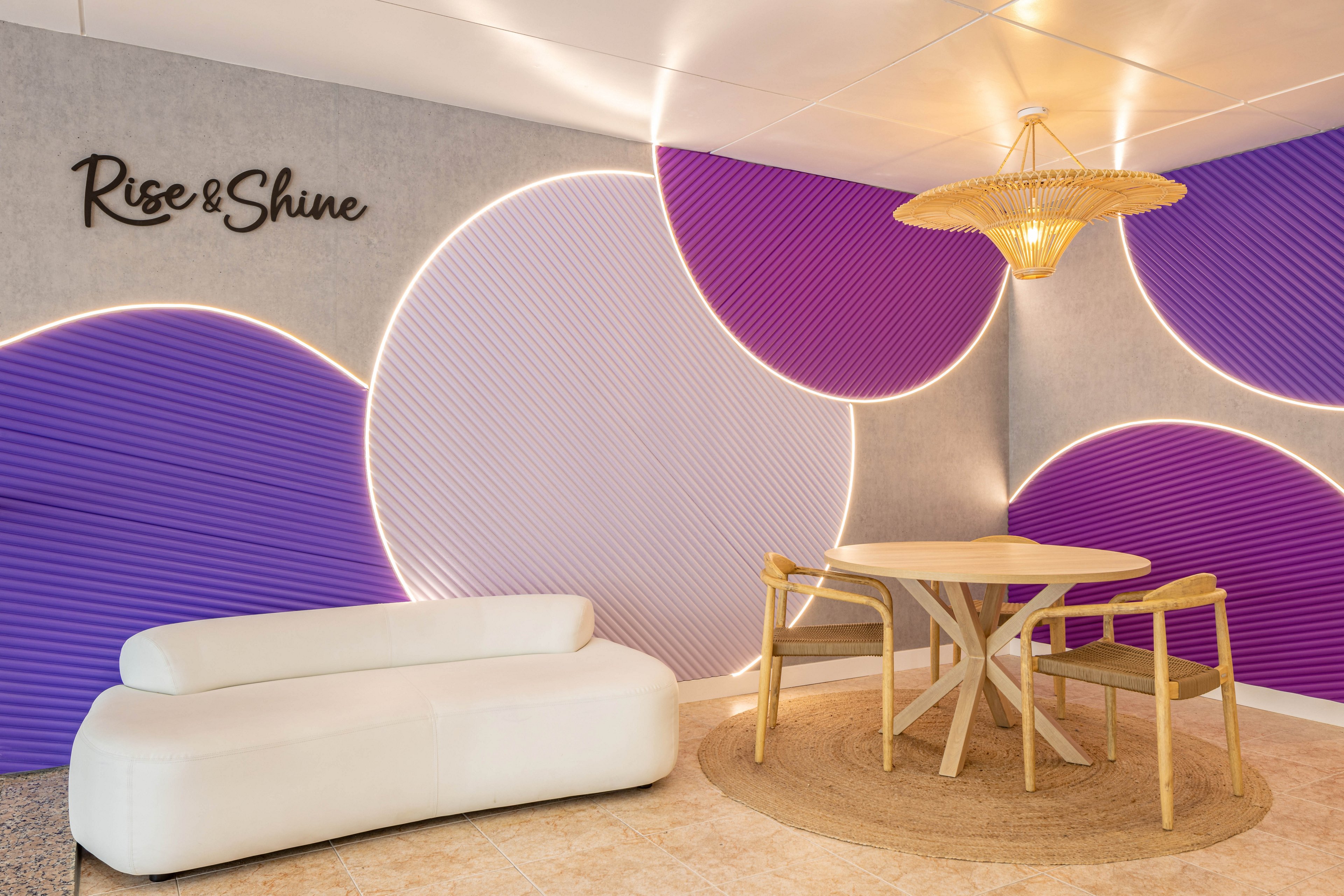 A contemporary lounge area features a white sofa and a round wooden table with matching chairs, set atop a circular rug. The wall displays bold purple geometric shapes illuminated by LED lighting, with the phrase 'Rise & Shine' clearly visible. A woven pendant lamp adds warmth to the stylish, inviting space.