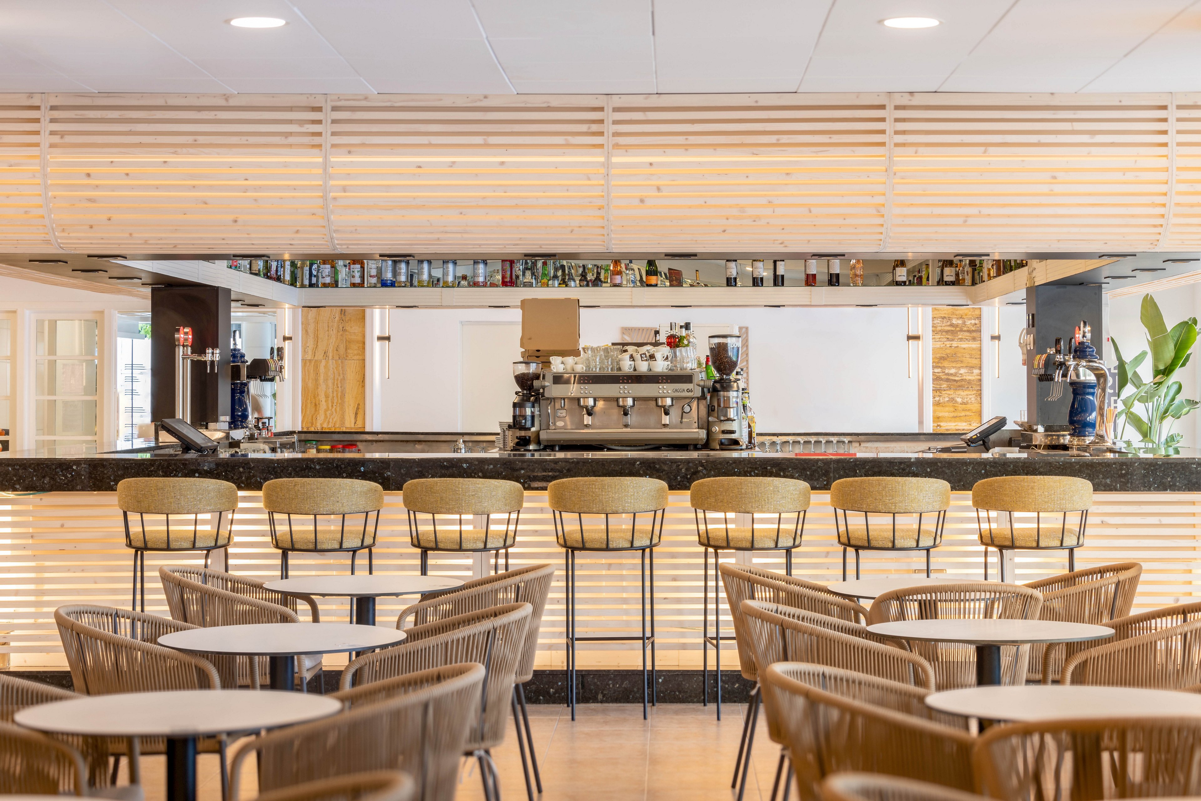 The image shows a contemporary café bar with a sleek counter and high stools arranged neatly. The setting is bright and airy, featuring light wood paneling and minimalist décor. Tables and chairs are placed in the foreground, while a coffee machine and bottles line the back counter. The overall mood is inviting and modern, with neutral tones and clean lines.