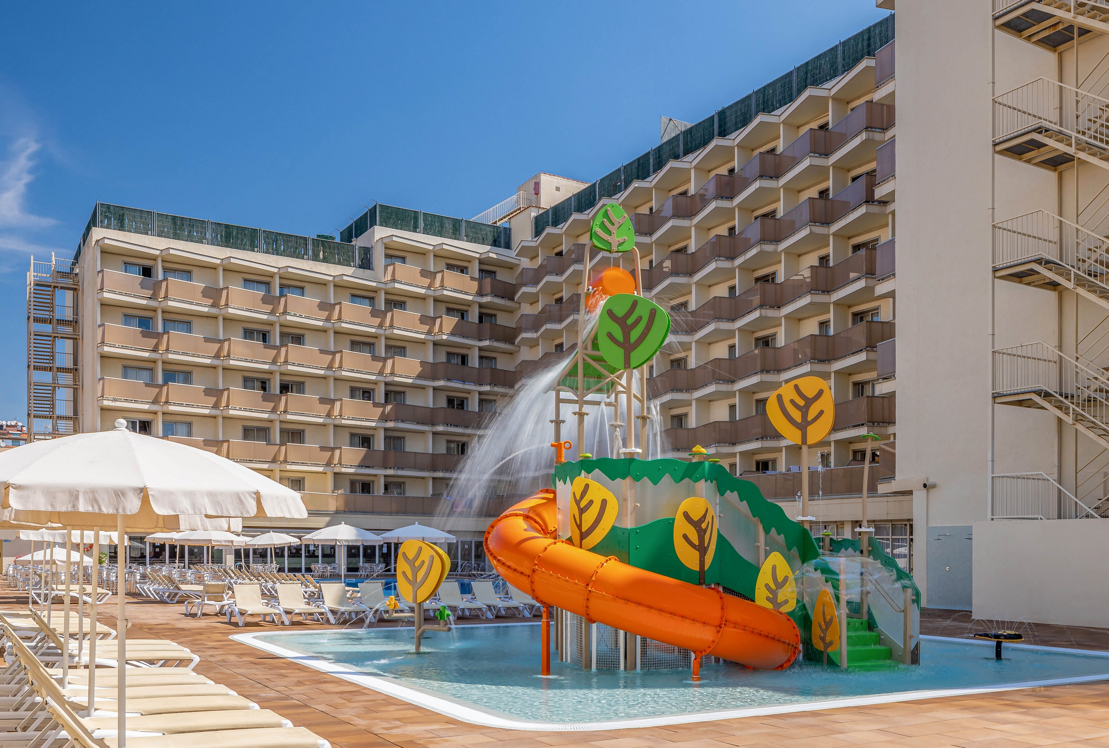 A vibrant outdoor pool area is shown in front of a multi-story hotel building. The main feature is a playful orange water slide surrounded by decorative green and yellow leaf motifs, with water spraying from above. The scene is set under a clear blue sky, with empty lounge chairs and umbrellas lining the poolside, creating a bright and inviting atmosphere.