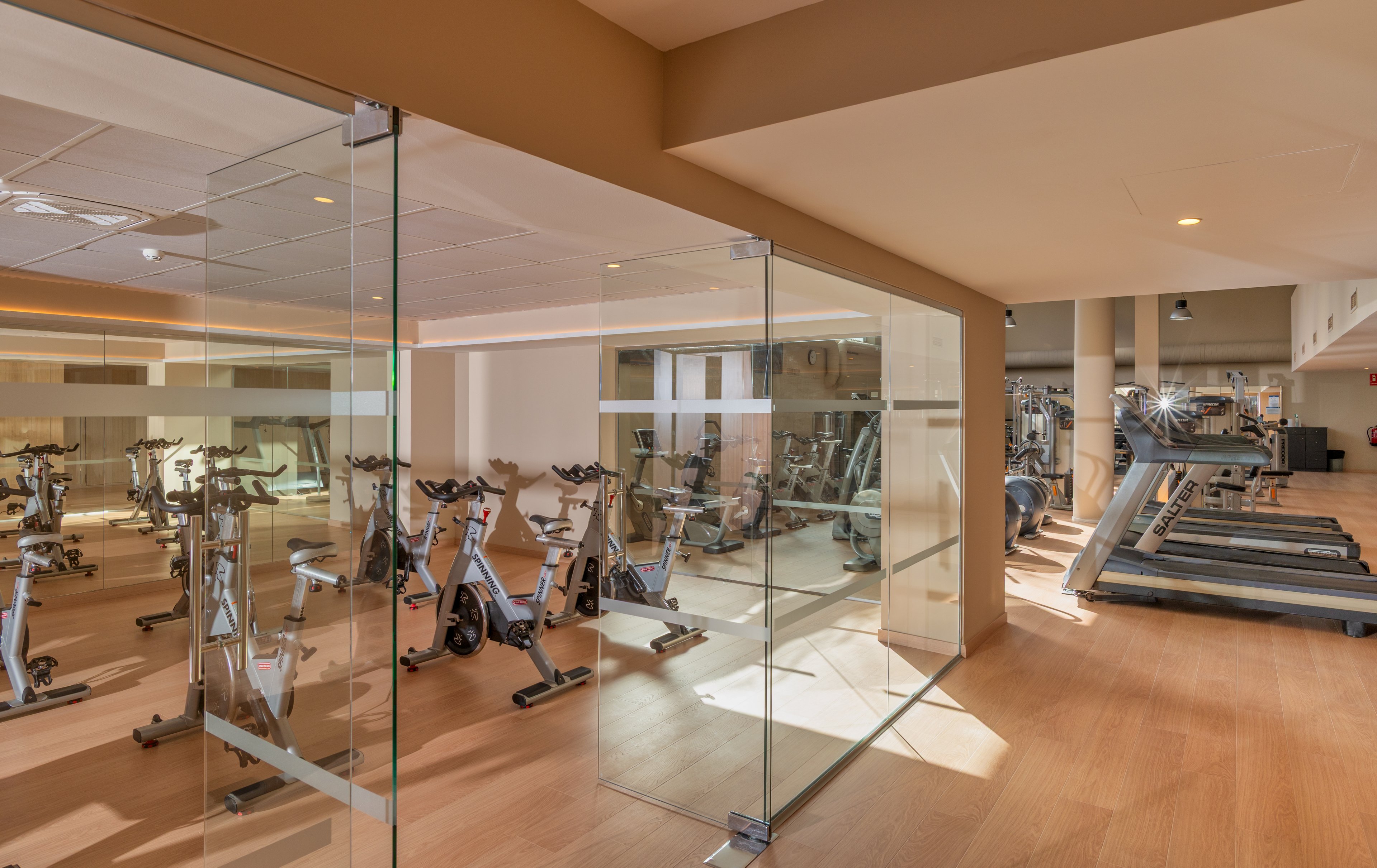 A contemporary gym interior features rows of stationary bikes and treadmills arranged neatly on a light wooden floor. The workout area is enclosed by clear glass walls, creating an open and spacious feel. Warm ceiling lights and neutral beige tones give the room a clean, inviting atmosphere. The space appears empty, ready for guests to begin their exercise routines.