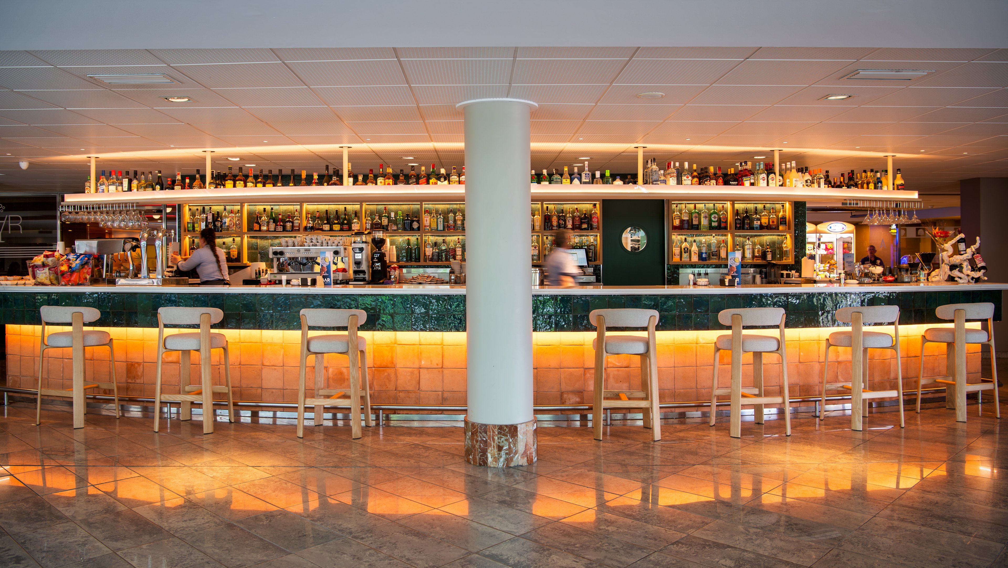 Wide view of a contemporary bar with a long counter and high stools arranged in a row. The back wall is filled with shelves of bottles and glassware, creating a colorful display behind the bar. Warm under-counter lighting reflects on the polished floor, giving the space a welcoming glow. A few bartenders work behind the counter, adding a sense of activity to the otherwise calm, modern interior.