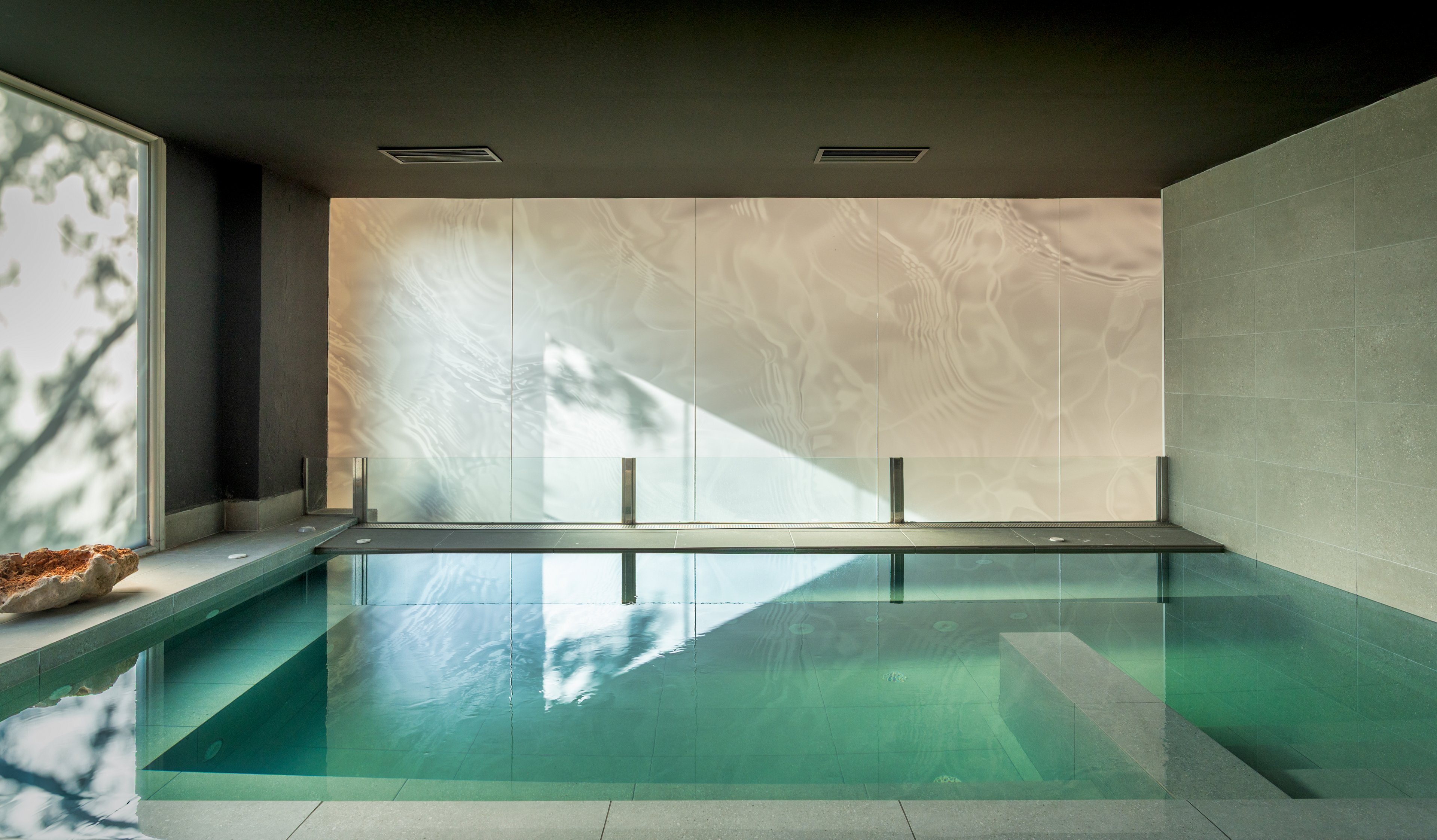 A serene indoor pool is set within a minimalist architectural space. Soft natural light streams through a large window, casting reflections on the pale textured wall. Clean lines, smooth stone surfaces, and muted colors create a calm, spa-like atmosphere. The still water and uncluttered design emphasize relaxation and modern luxury.