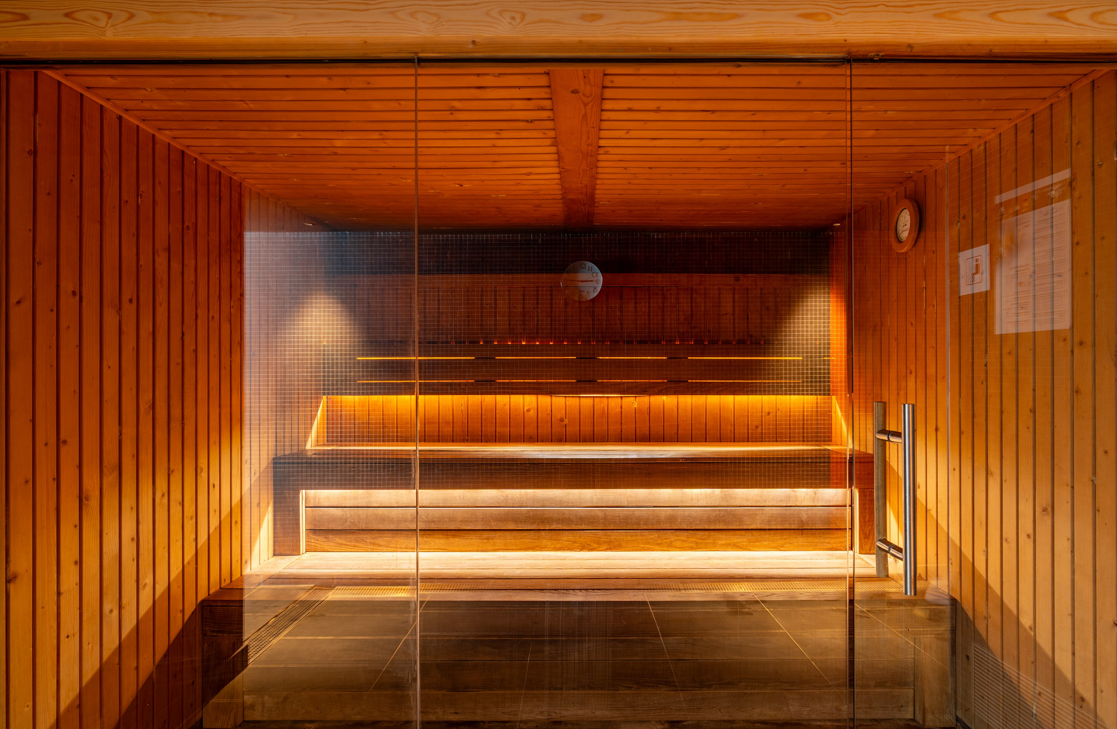 A modern wooden sauna interior is viewed through a glass door. Tiered wooden benches line the back wall, illuminated by soft, warm strip lighting. The walls and ceiling are clad in vertical wooden panels, creating a cozy and inviting atmosphere. A small round thermometer and a posted notice are visible on the right wall, emphasizing the functional spa setting.