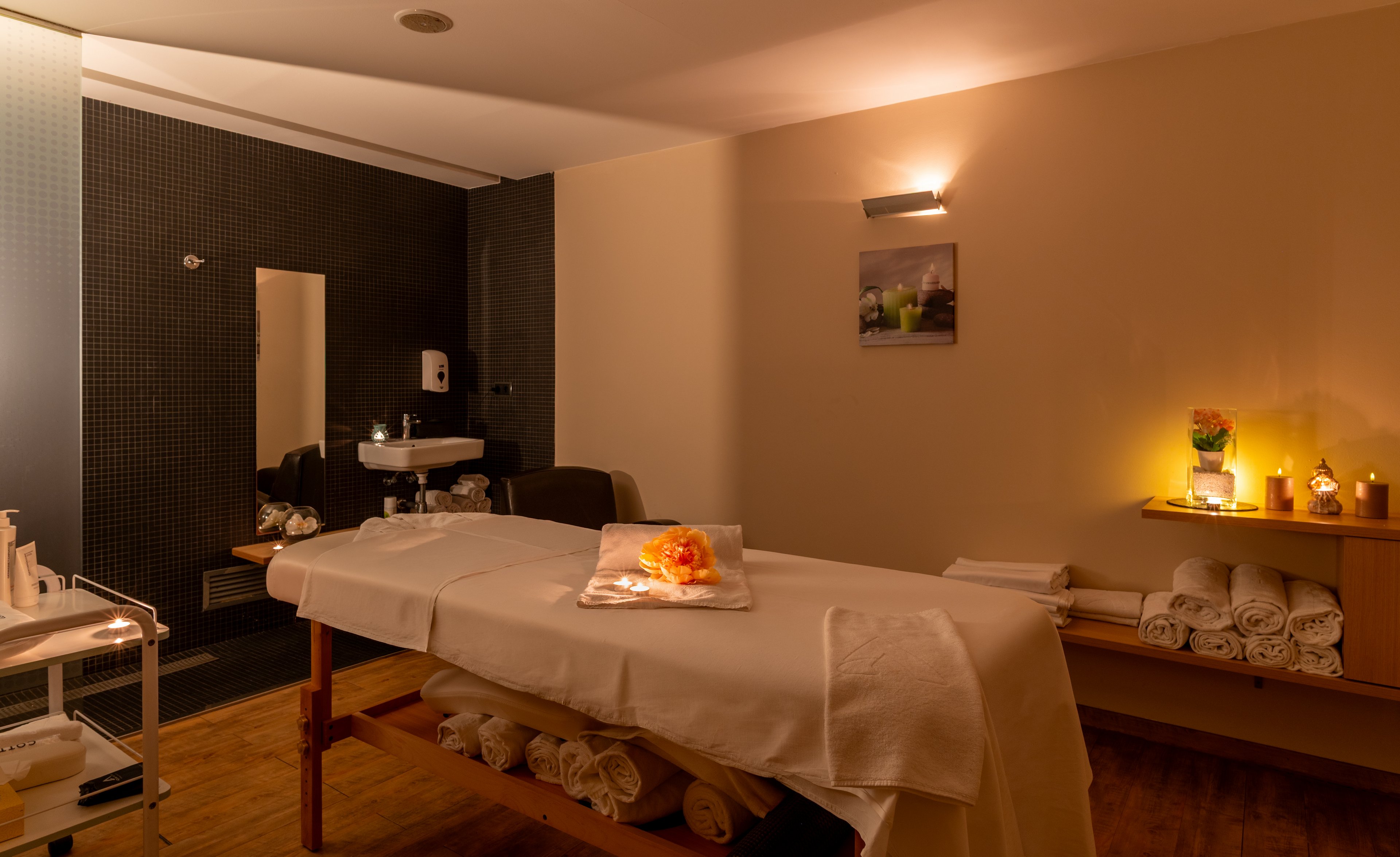 A neatly prepared massage table stands in a softly lit spa treatment room. Rolled white towels are stacked on shelves and under the table, suggesting readiness for clients. Warm candlelight and a small flower arrangement create a relaxing, intimate atmosphere. A sink and dark-tiled shower area are visible in the background, indicating a fully equipped wellness space.