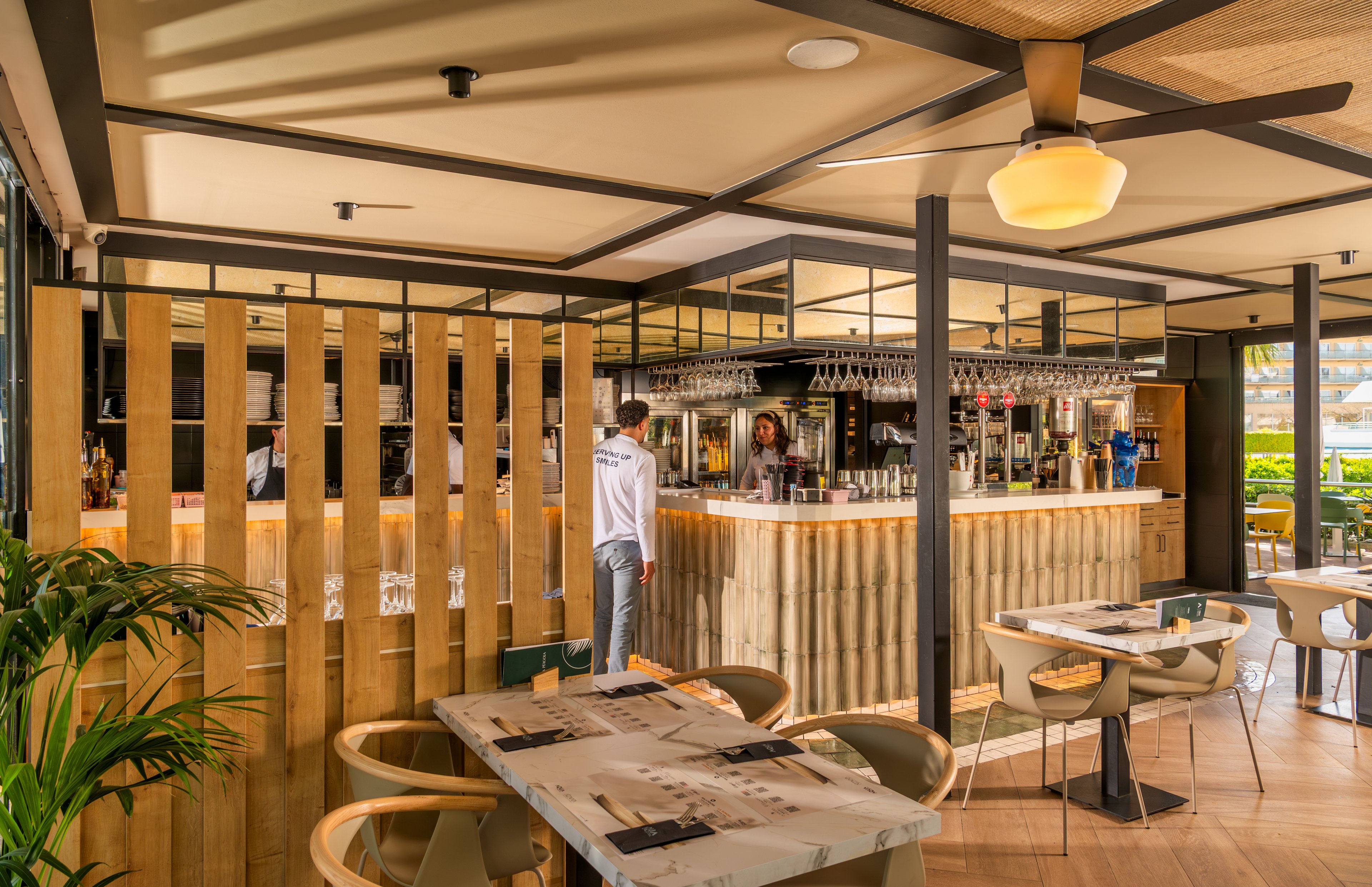 Contemporary restaurant interior featuring a central bar area with wooden paneling and mirrored shelving. A staff member stands behind the bar, surrounded by hanging glassware and bottles. Light wood tables and curved chairs are neatly arranged in the foreground, creating an inviting dining space. Large windows and indoor plants add natural light and greenery to the modern, relaxed atmosphere.