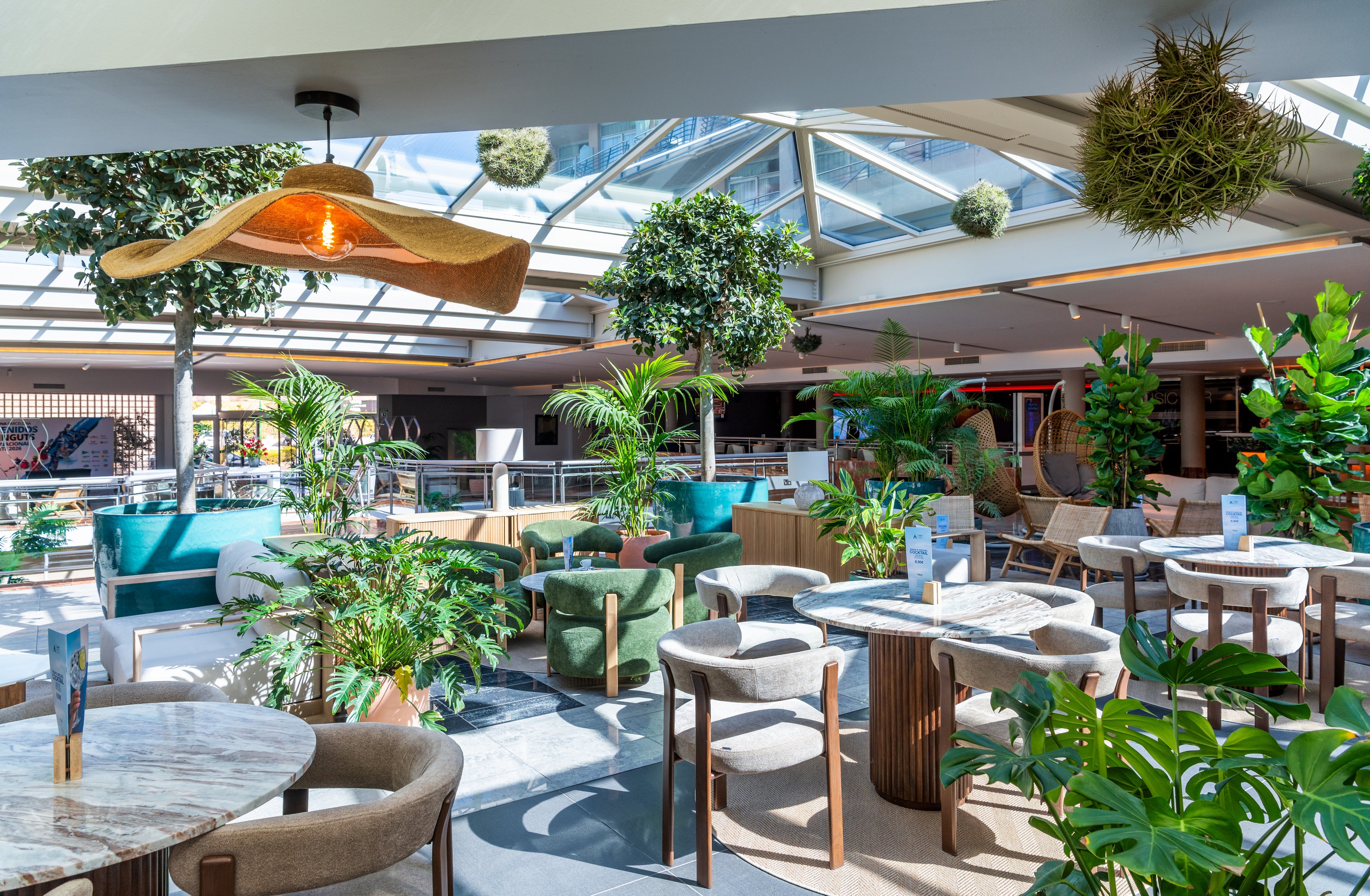 A spacious contemporary cafe interior is filled with natural light from a large glass skylight. Various tables and upholstered chairs are arranged in cozy seating areas, surrounded by numerous potted plants and hanging greenery. A sculptural woven pendant lamp glows warmly above the space, adding contrast to the cool daylight. The overall atmosphere is relaxed and inviting, blending modern design with a vibrant indoor garden feel.