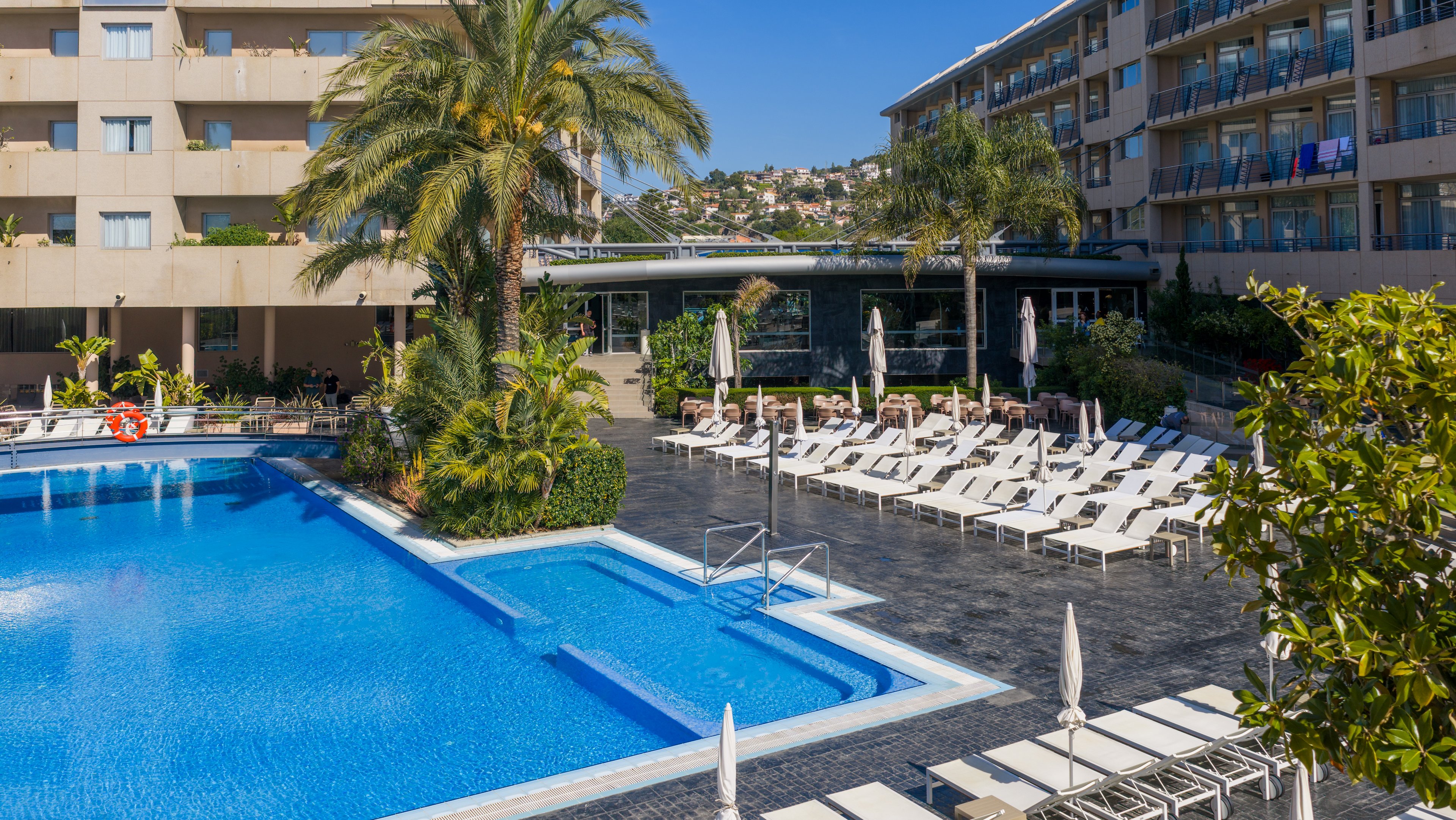 A large outdoor swimming pool sits at the center of a modern hotel courtyard. Numerous white sun loungers and umbrellas line the dark tiled pool deck, creating a resort-style relaxation area. Tall palm trees and lush greenery add a tropical touch to the urban hotel buildings surrounding the pool. Clear blue sky and distant hillside houses suggest a warm, sunny vacation destination.