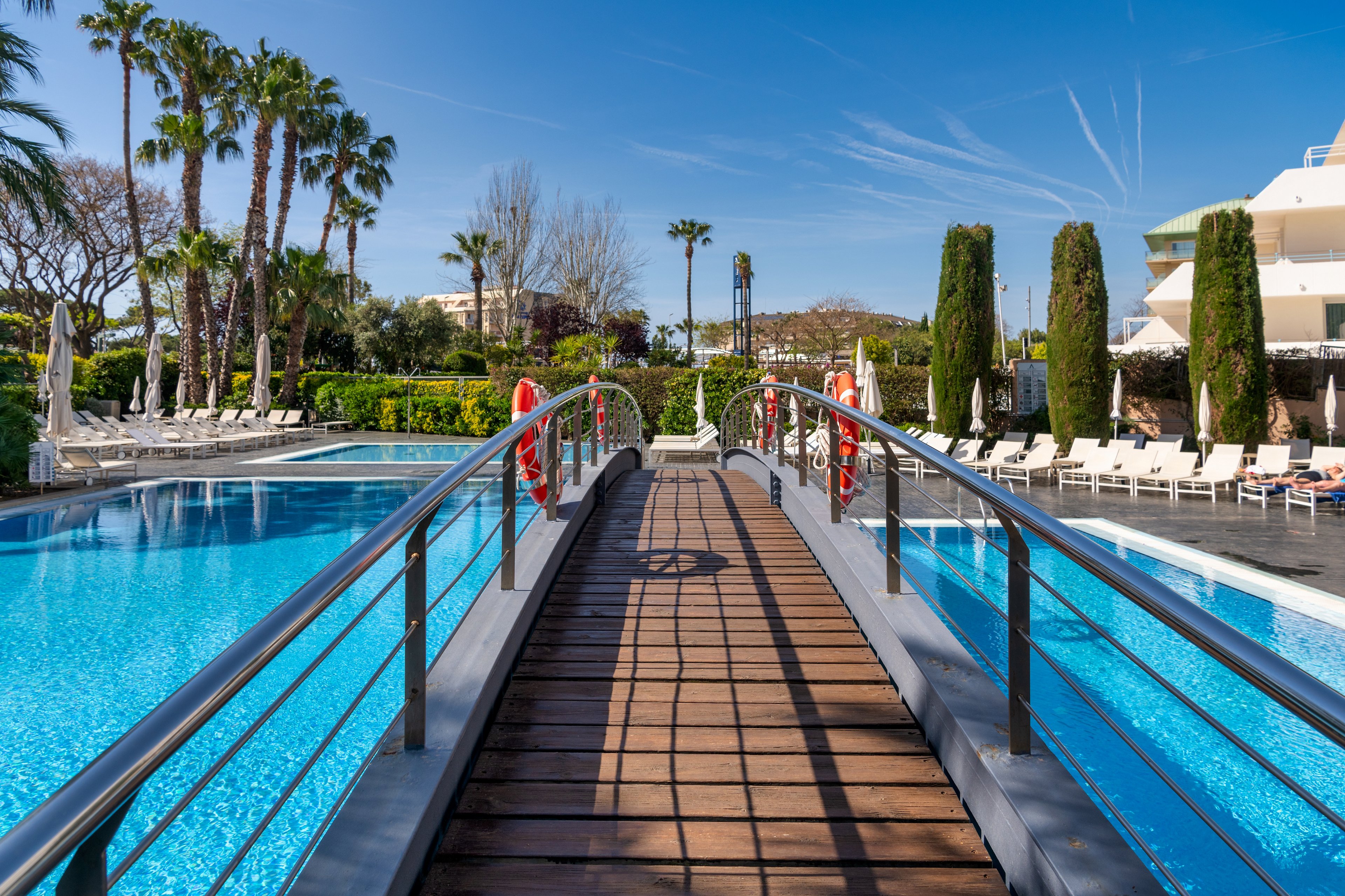 A wooden footbridge with metal railings spans a large, clear blue swimming pool at a sunny resort. Tall palm trees and manicured hedges surround the pool area, creating a lush, tropical atmosphere. White lounge chairs line the poolside under a bright blue sky with thin clouds and jet trails. Modern resort buildings are visible in the background, emphasizing a relaxing vacation setting.