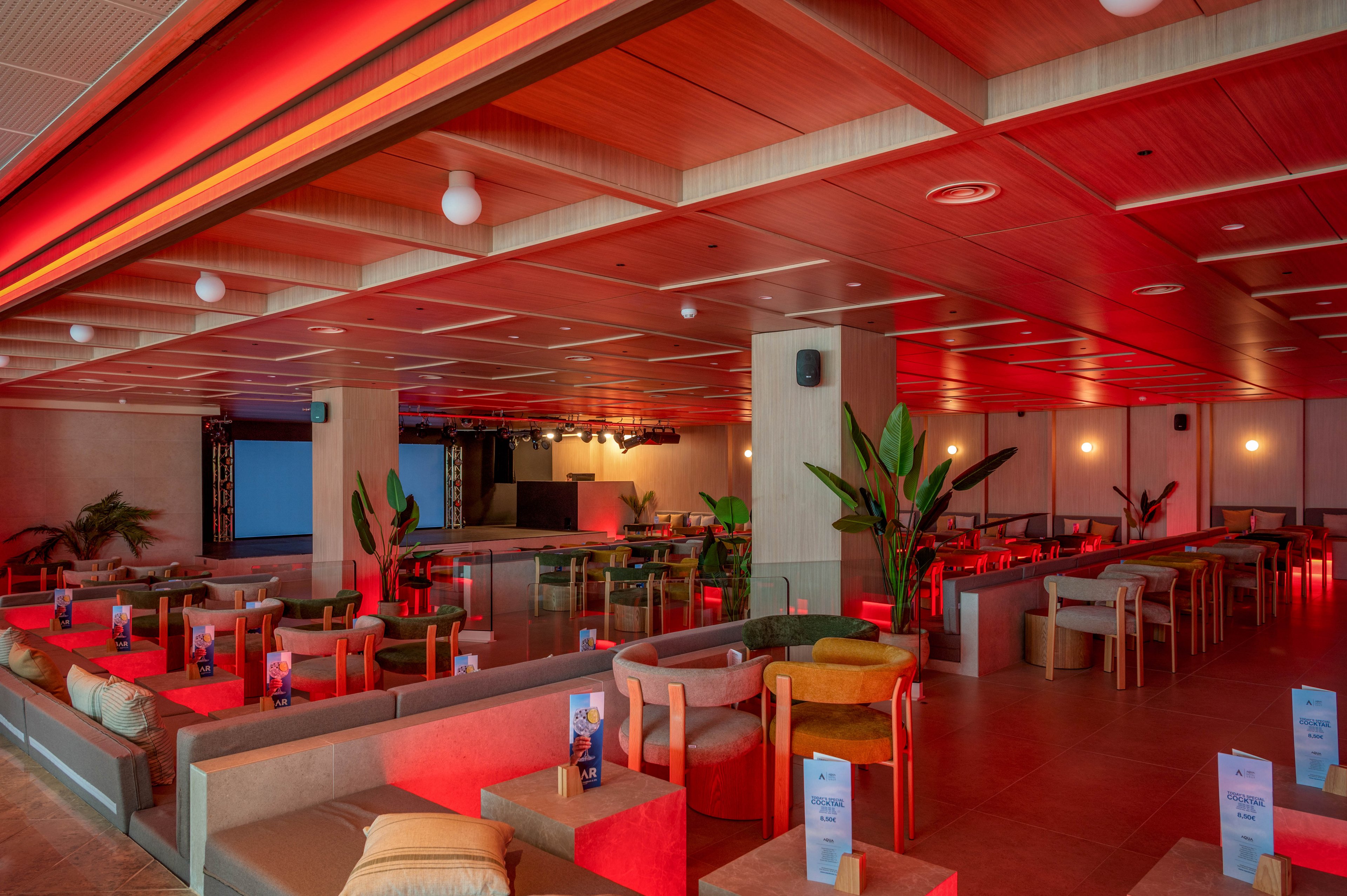A spacious modern lounge bar features low sofas, armchairs, and small tables arranged in neat rows. Warm red ambient lighting washes over the wooden ceiling and neutral-toned furniture, creating a stylish evening atmosphere. Potted plants add greenery between seating areas, while table tents and menus stand upright on each table. A stage with a large screen and DJ booth is visible in the background, suggesting the venue is prepared for events or performances.