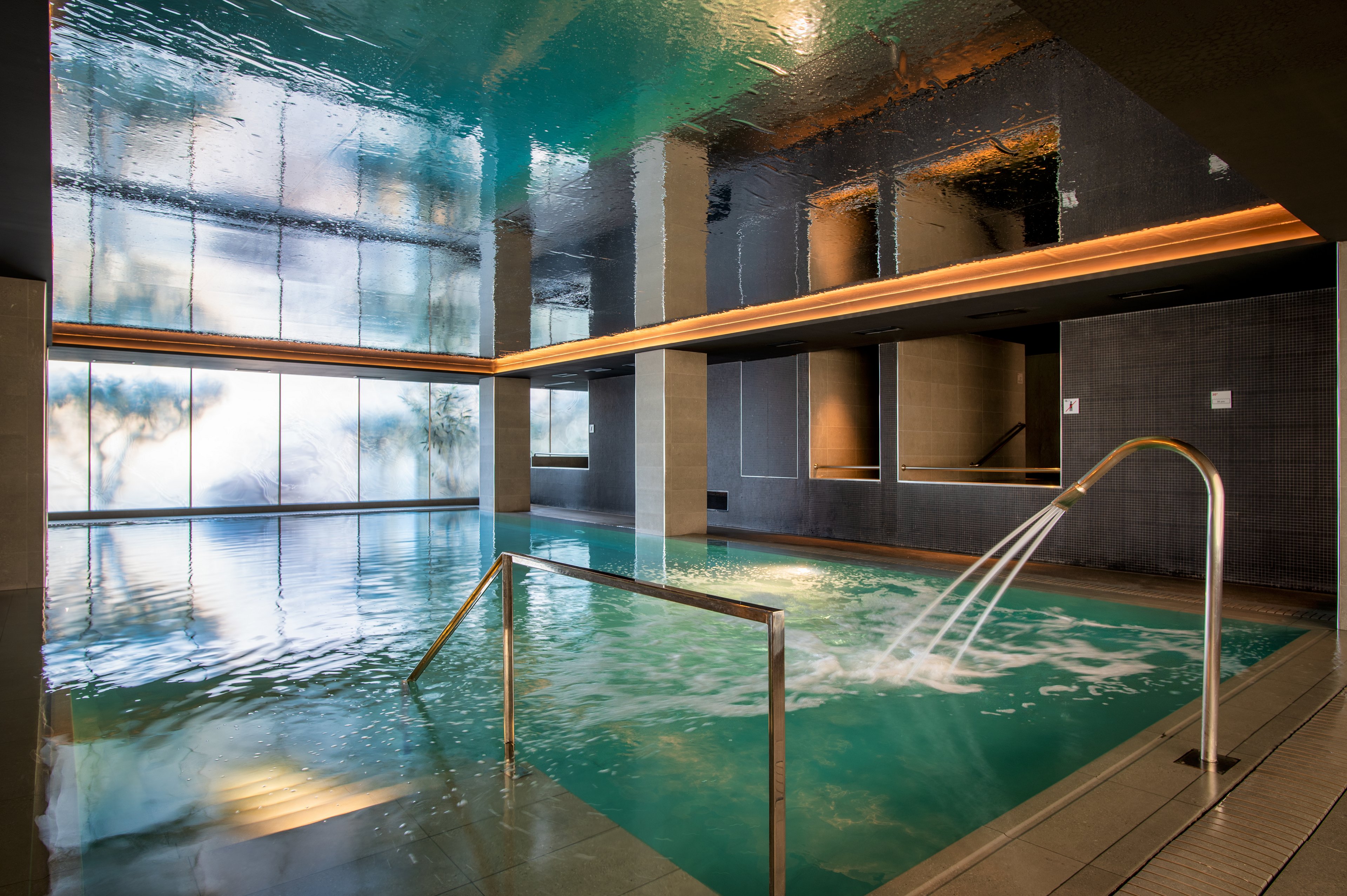 A contemporary indoor spa pool features stainless steel handrails and arched water jets flowing into turquoise water. The space is enclosed by large reflective windows and dark tiled walls, creating a sleek, luxurious atmosphere. Warm recessed lighting runs along the ceiling edge, contrasting with the cool tones of the pool. The scene is calm and empty, emphasizing relaxation and wellness.