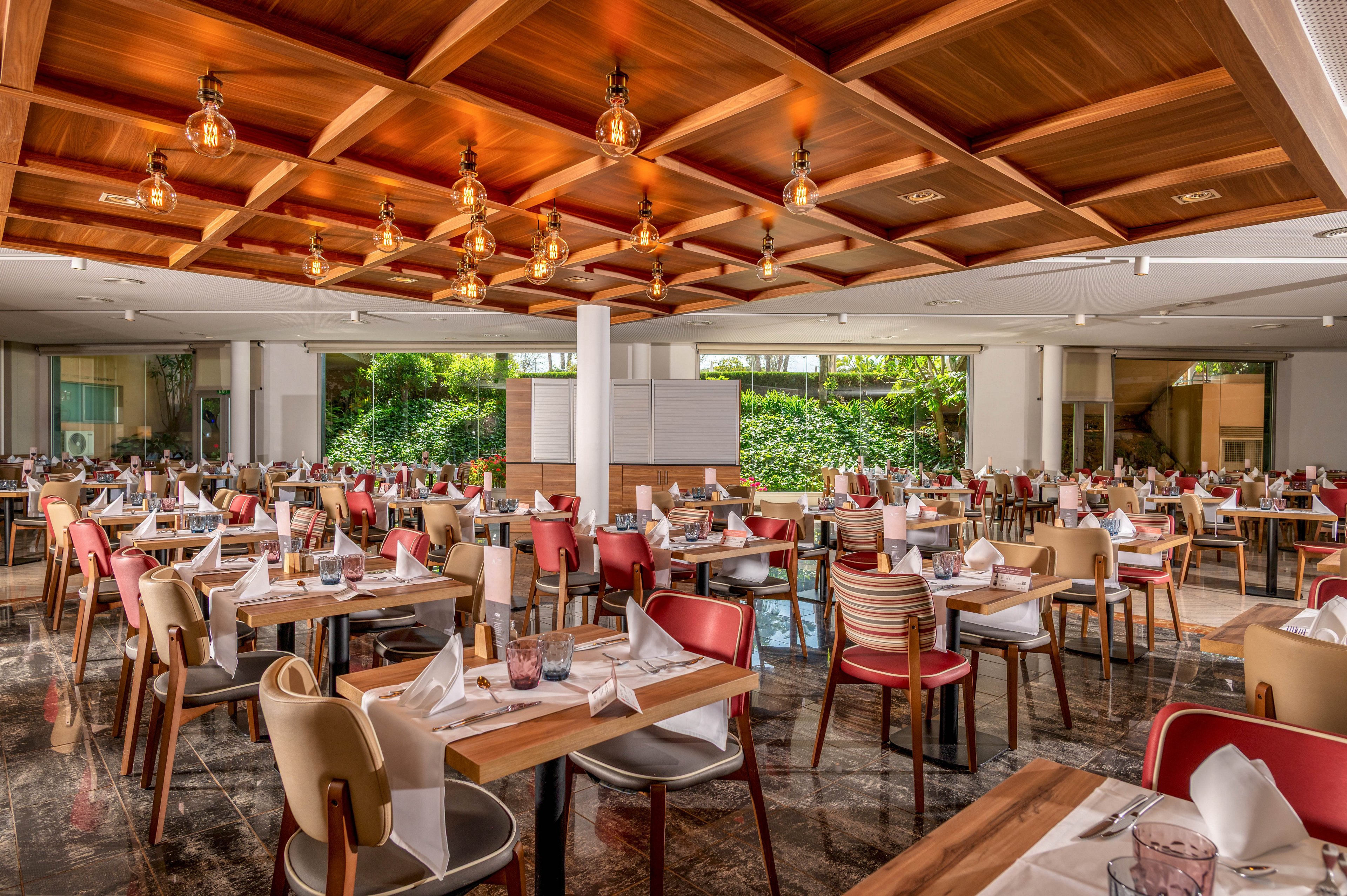 Spacious restaurant dining room with neatly arranged wooden tables and chairs. Each table is set with white napkins, glassware, and cutlery, ready for guests. Warm pendant lights hang from a wooden coffered ceiling, casting a cozy glow over the polished floor. Large windows or openings reveal lush greenery outside, adding a fresh, inviting atmosphere to the modern space.