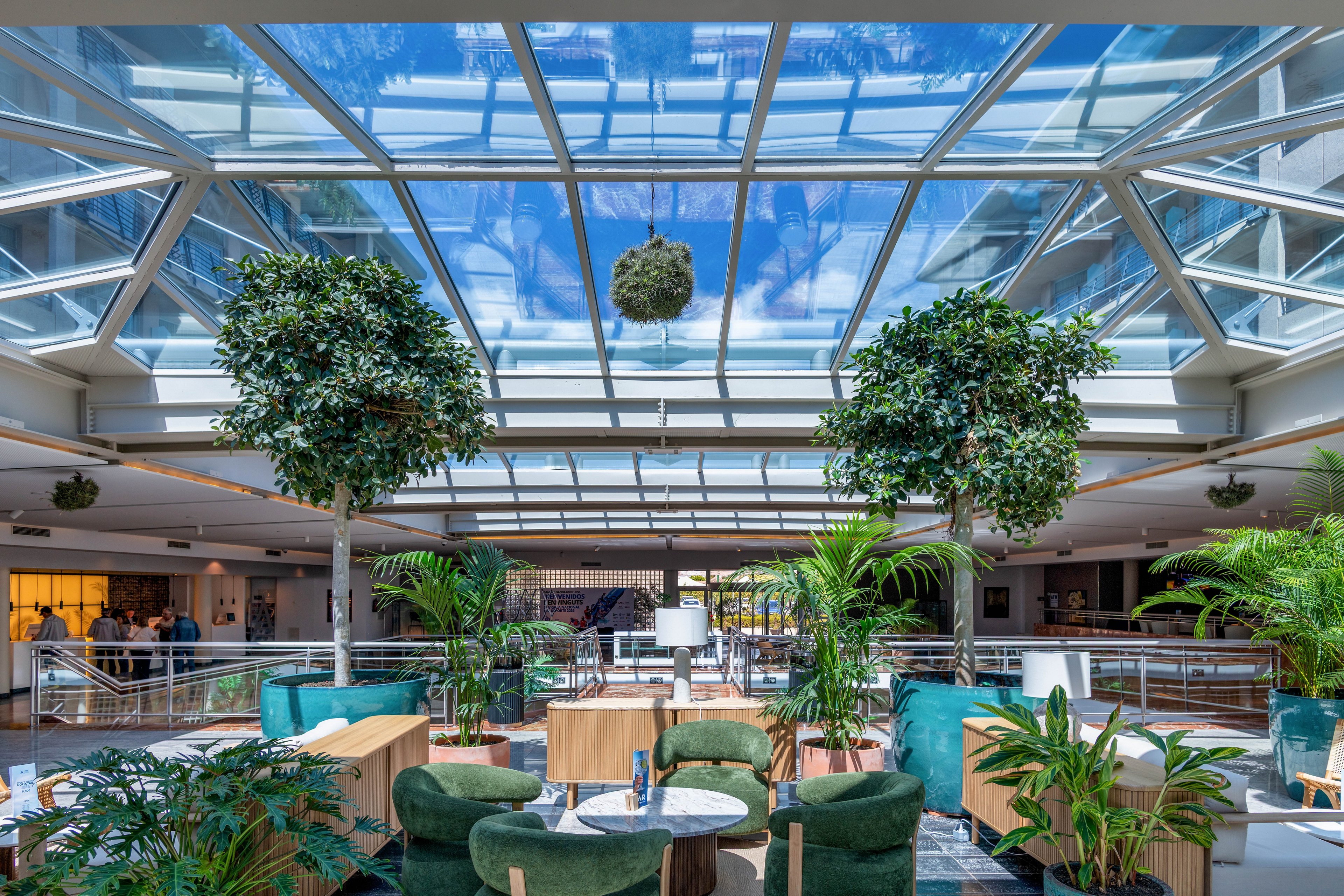 A spacious indoor atrium lounge features abundant greenery and contemporary seating beneath a large glass skylight. Tall trees and potted plants surround plush green chairs and wooden tables arranged for casual meetings or relaxation. Natural daylight floods the space through the geometric glass ceiling, creating a bright and airy atmosphere. People can be seen in the background near a corridor, adding a subtle sense of activity to the tranquil setting.