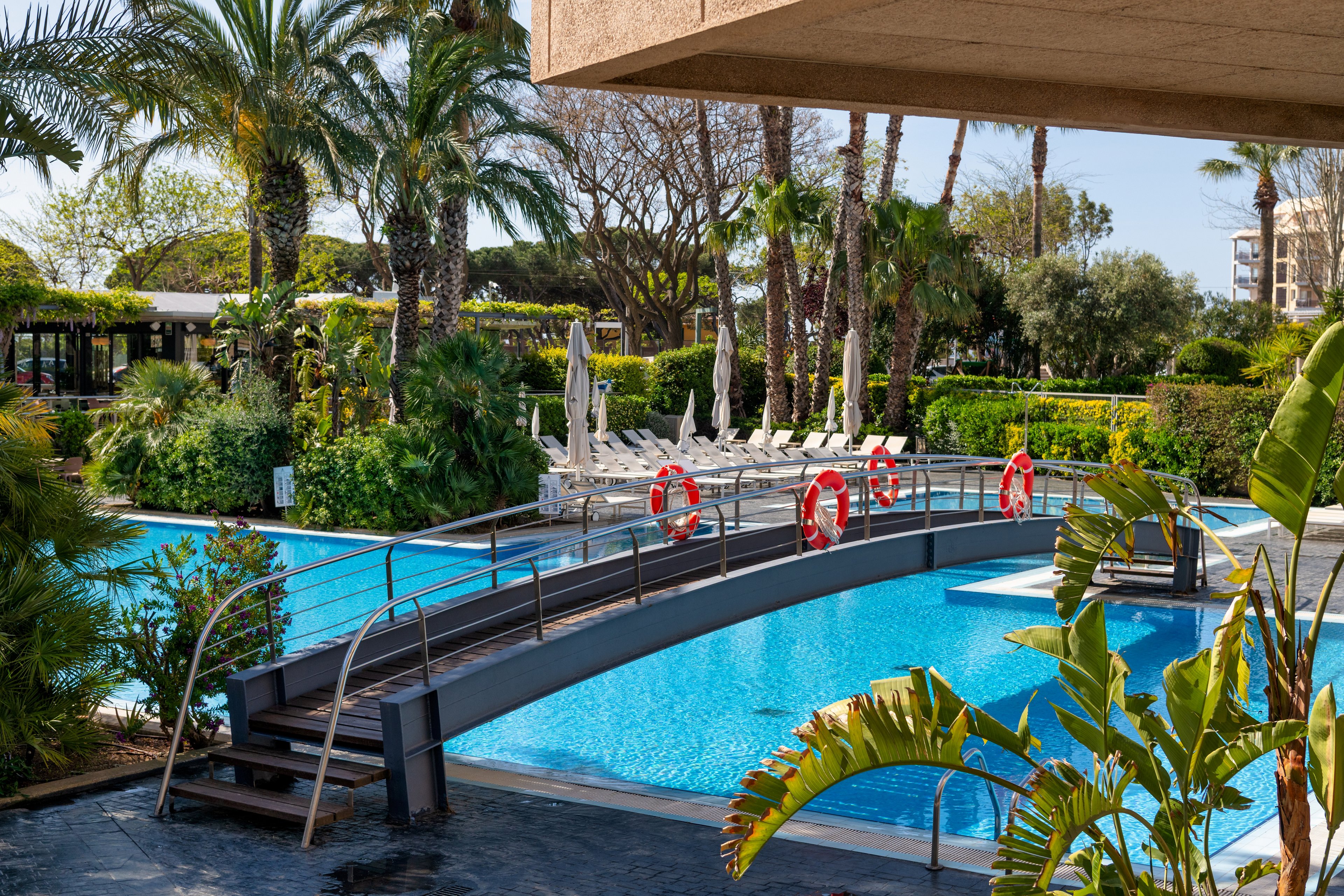 An outdoor resort swimming pool features a curved metal footbridge crossing over bright blue water. White sun loungers and umbrellas are arranged in the background, suggesting a relaxing vacation setting. Palm trees and lush green plants surround the pool area, creating a tropical atmosphere. Red life rings hang along the bridge railing, adding a vivid accent and safety detail to the tranquil scene.
