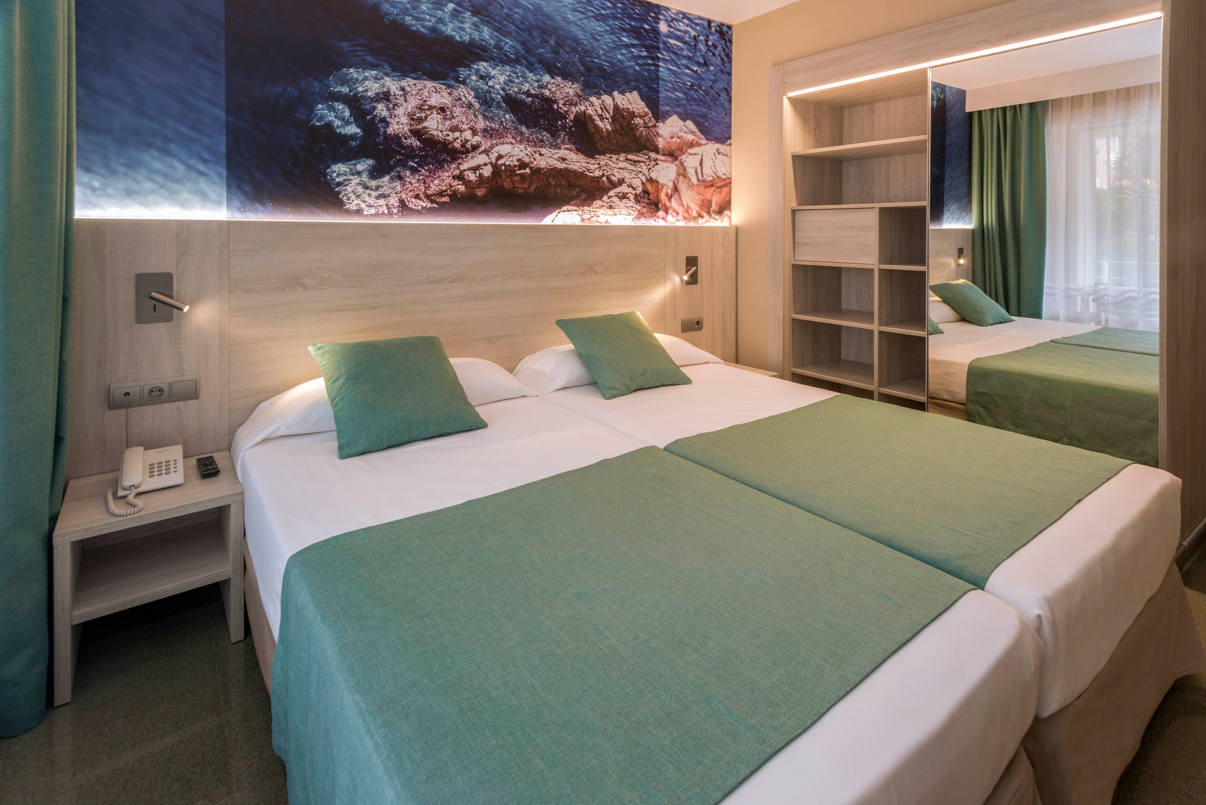 Contemporary hotel room featuring two neatly made twin beds with coordinated green bedding and pillows. A large wall mural above the headboard shows a rocky coastline under clear blue water, adding a coastal theme to the space. Built-in shelving, a bedside table with a telephone, and a mirrored wardrobe provide functional storage. Soft lighting and floor-to-ceiling curtains create a calm, inviting atmosphere.