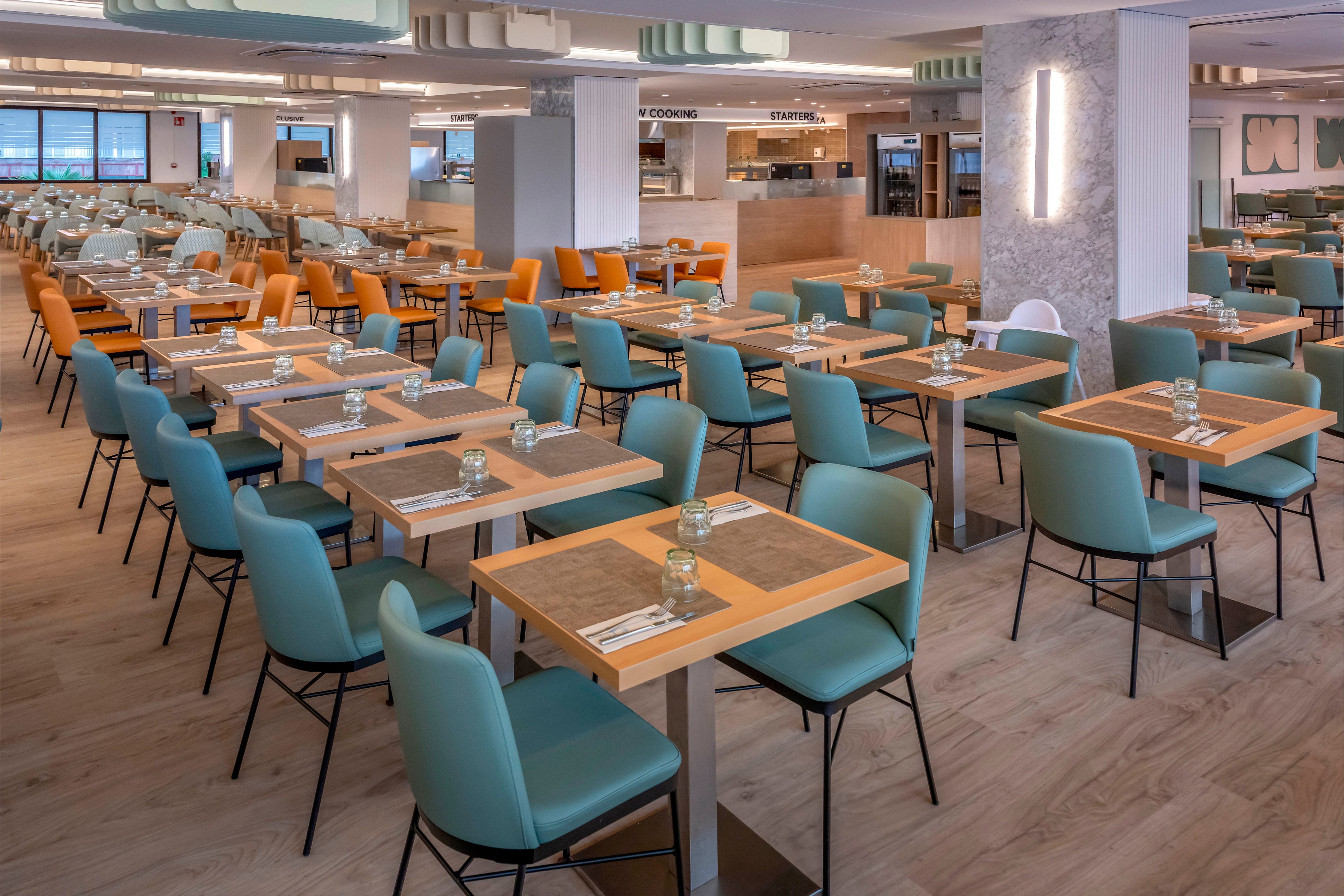 A spacious, contemporary cafeteria is shown with rows of neatly arranged wooden tables and pastel-colored chairs. Each table is set with placemats, cutlery, and small glass containers, ready for guests. The interior features light wood flooring, soft overhead lighting, and a clean, minimalist serving area in the background. Large windows on one side bring in natural light, enhancing the bright and welcoming atmosphere.