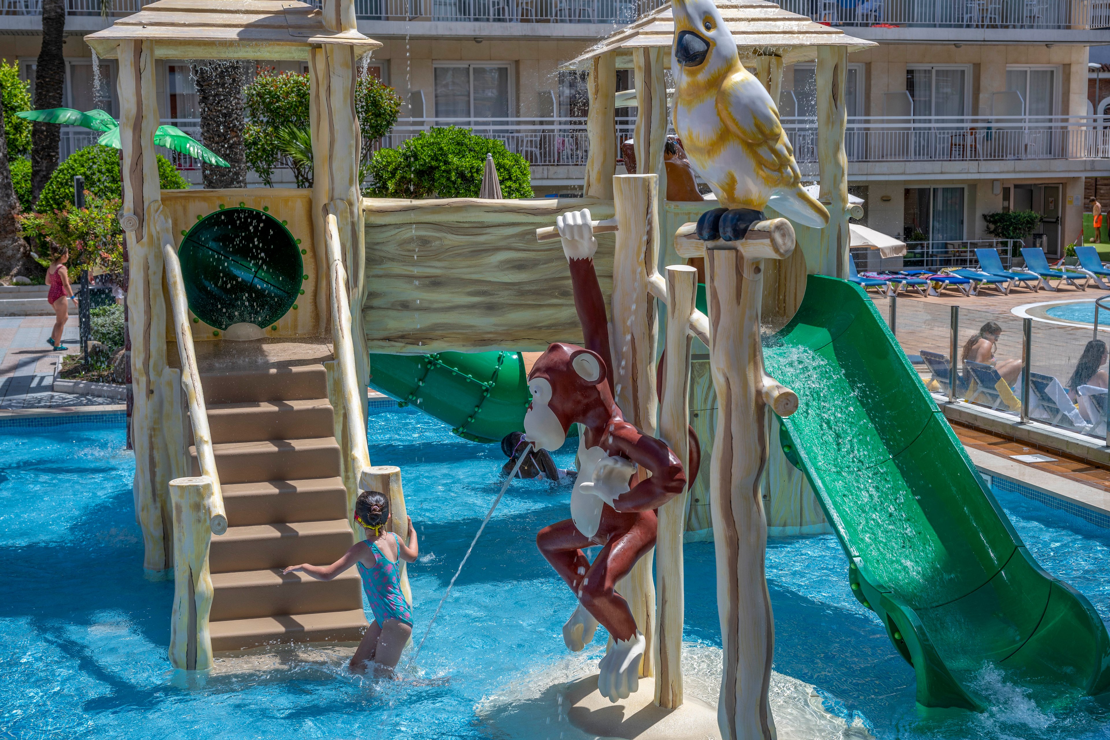 A child plays in a shallow outdoor pool at a themed water playground. The structure features a large green slide, wooden-style steps, and a hanging monkey figure. A big bird statue perches on top of the play equipment, while water splashes around the child. In the background, hotel balconies, sun loungers, and other guests suggest a vacation resort setting.