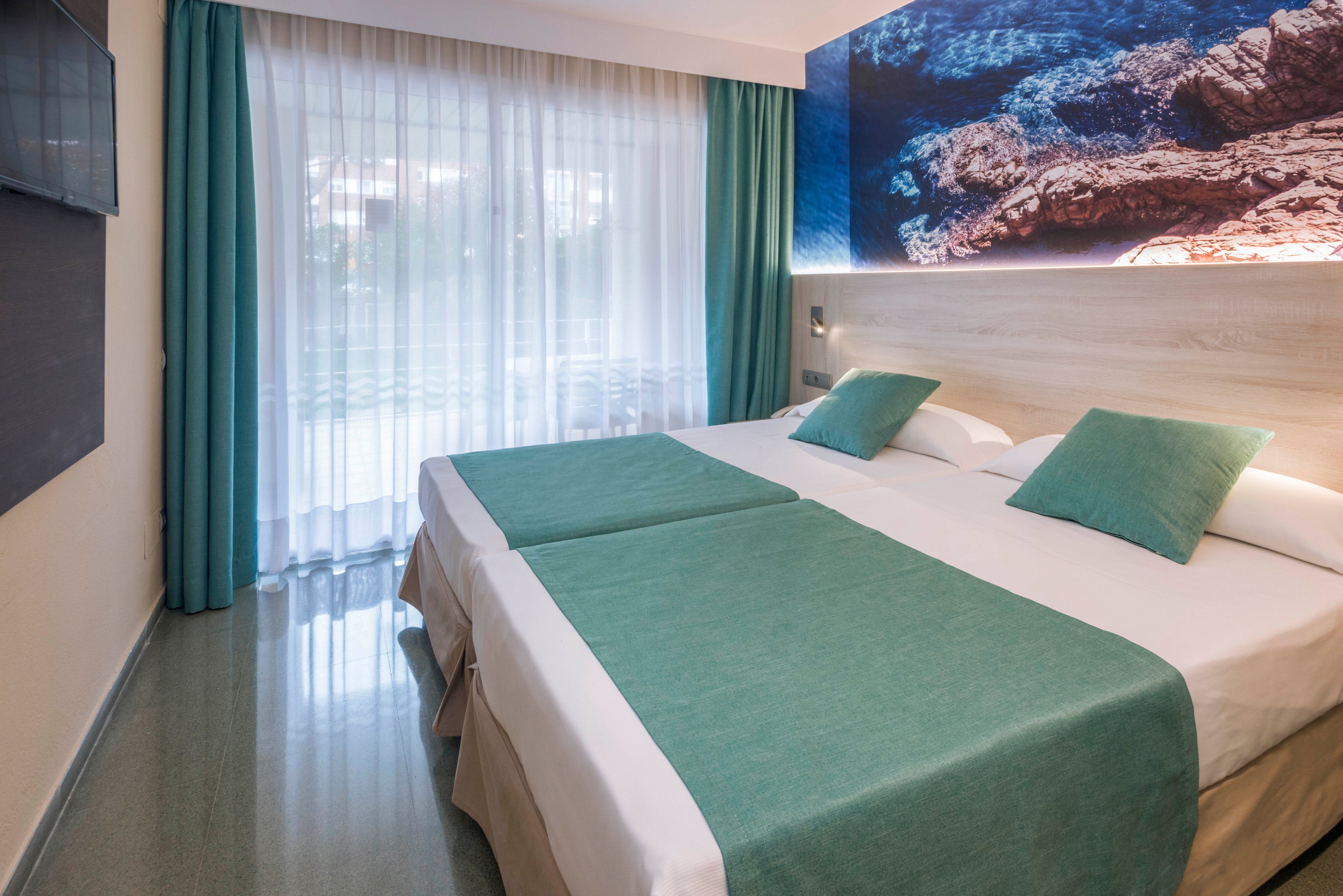Contemporary hotel room interior featuring two neatly made twin beds pushed together with teal bed runners and pillows. Large sheer curtains cover a window or balcony door, allowing soft natural light to fill the space. A wall-mounted artwork of a rocky coastline with blue water adds a coastal accent above the headboard. The room has a clean, minimalist design with cool tones and polished flooring, creating a calm and inviting atmosphere.