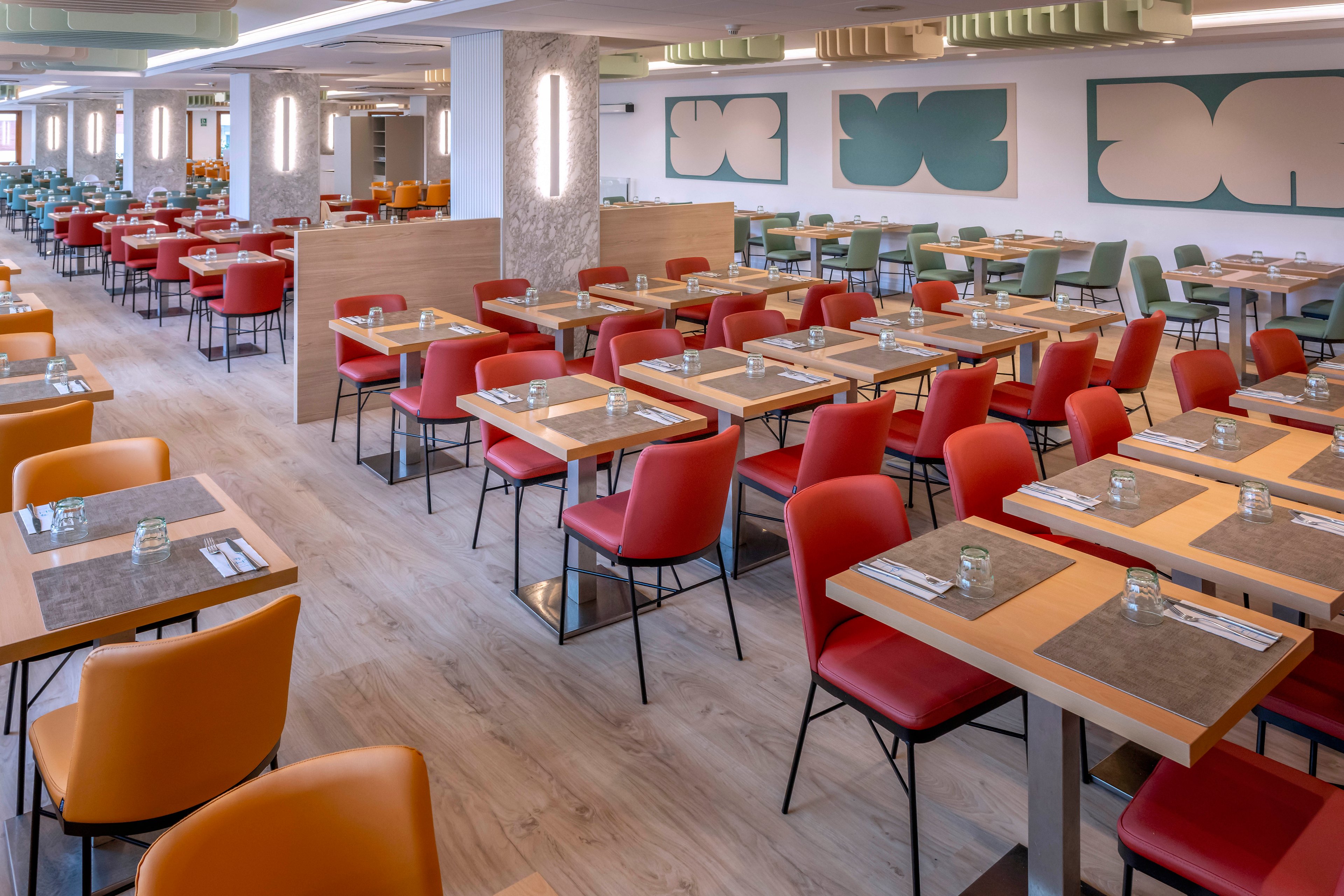 A spacious contemporary restaurant dining room is filled with neatly arranged tables and chairs. Red and orange upholstered chairs surround wooden tables set with glasses, placemats, and cutlery. The room features light wood flooring, soft ceiling lighting, and decorative wall panels with abstract shapes. No people are present, emphasizing the clean, orderly atmosphere and modern design.