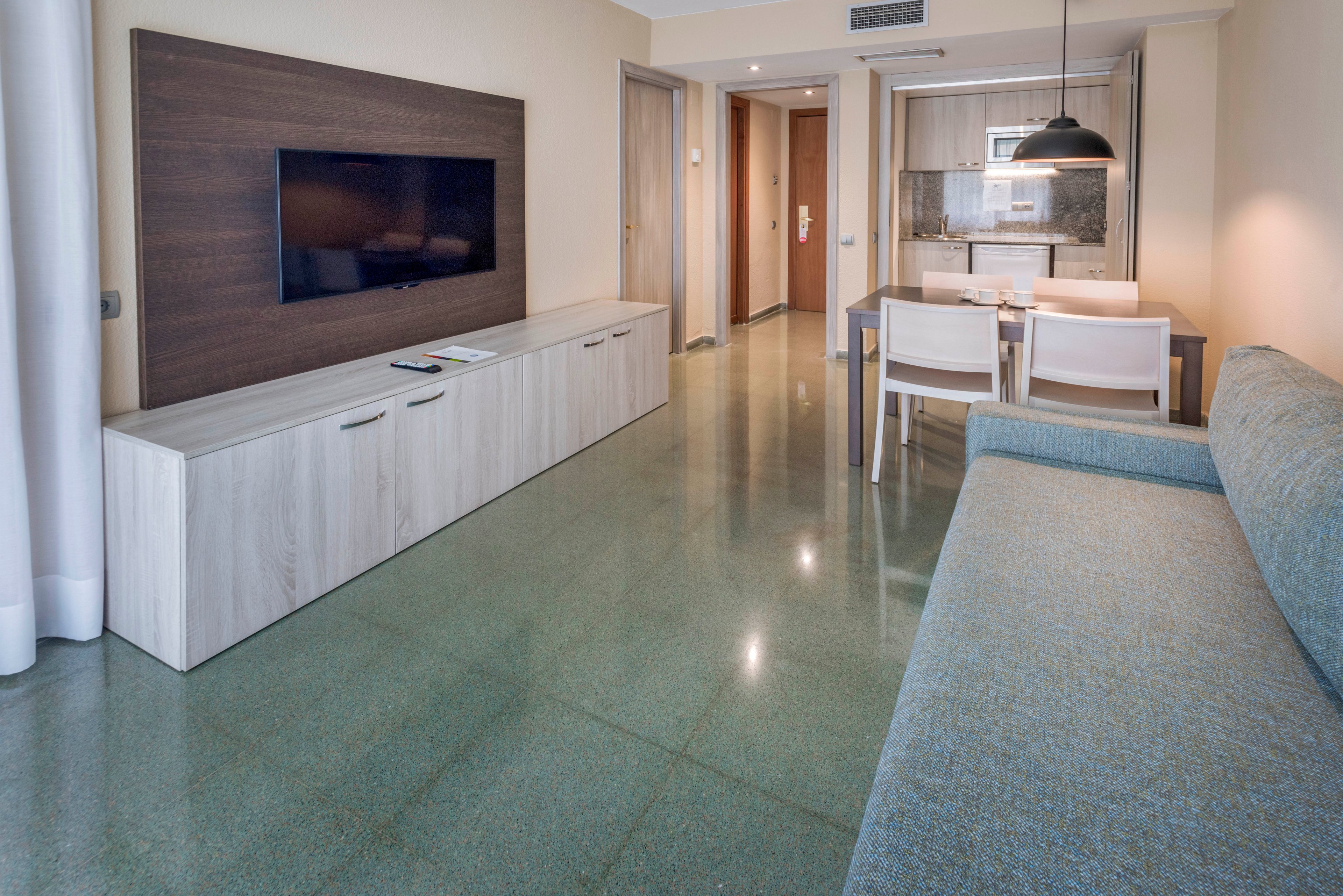 Contemporary hotel suite interior featuring a wall-mounted flat-screen television and long light-wood media console. A small dining table with two white chairs sits near a compact kitchenette in the background. A large sofa lines one side of the room, facing the television and console. The space is clean and minimalist, with polished green flooring and neutral-colored walls creating a calm, modern atmosphere.