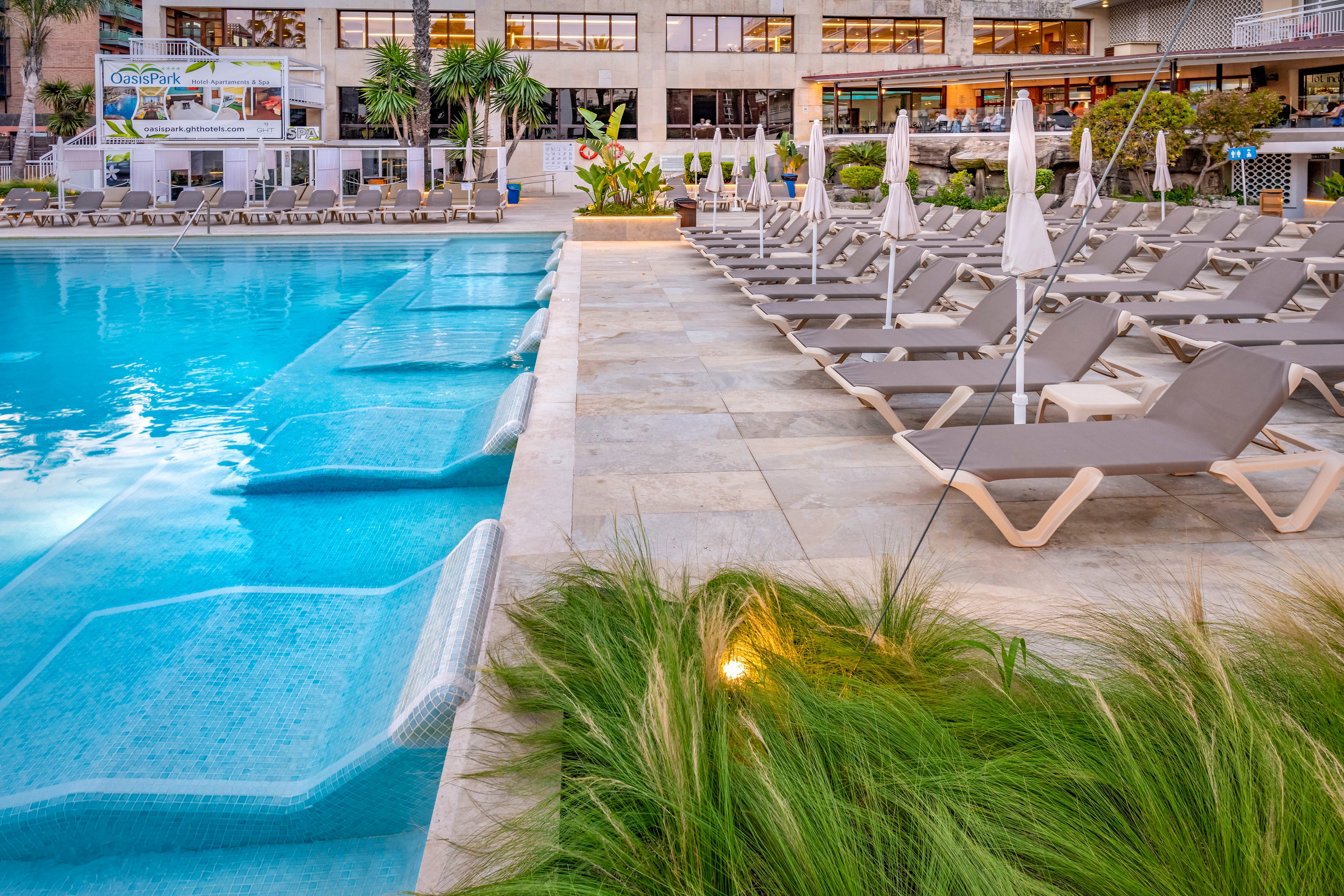 A contemporary resort swimming pool features built-in submerged loungers along the water’s edge. Rows of empty sun loungers with closed umbrellas line the stone pool deck, suggesting an early or quiet time of day. Lush ornamental grasses and tropical plants soften the hardscape and add greenery to the scene. In the background, a multi-story hotel building with large windows and balconies overlooks the pool area.
