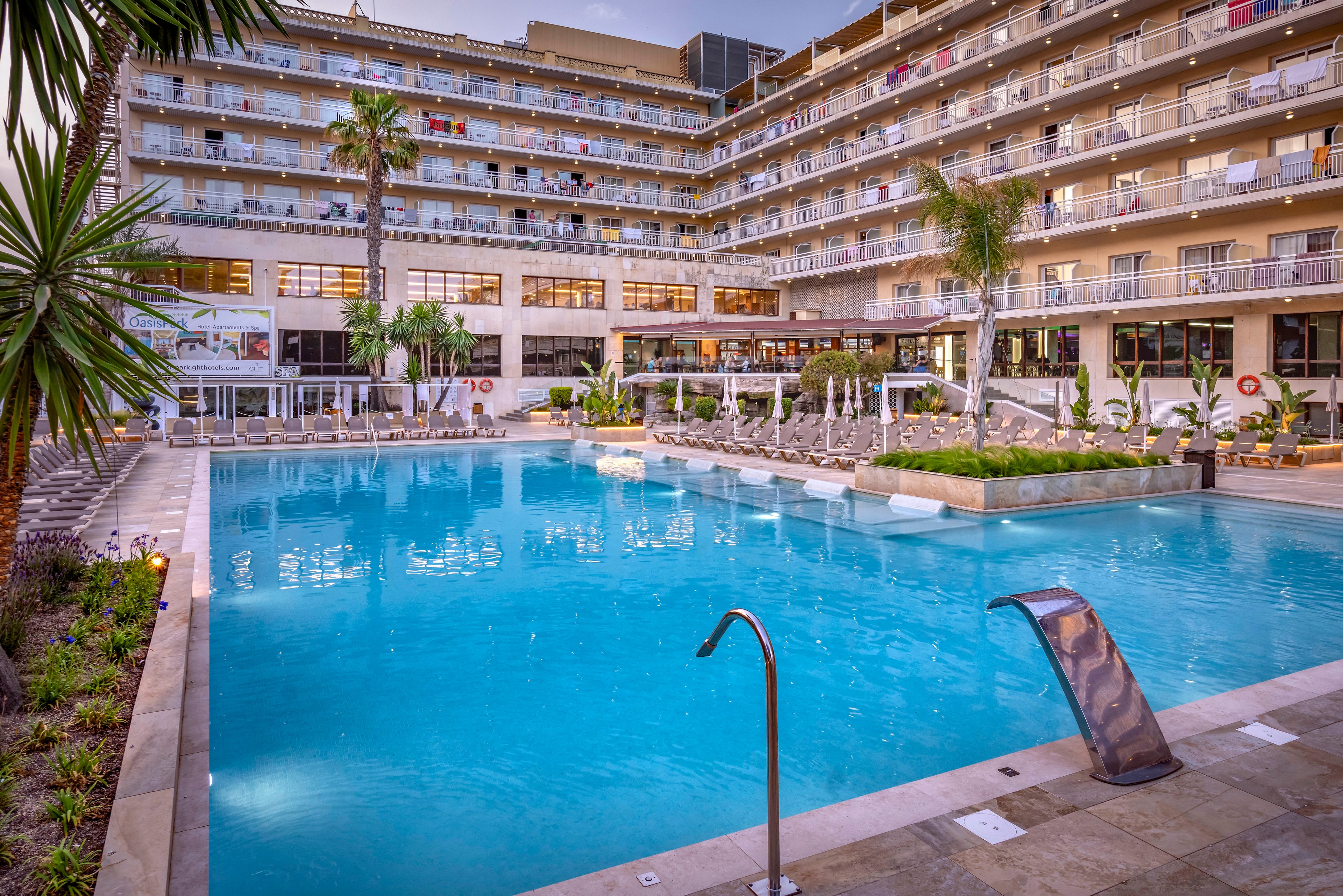 A contemporary multi-story resort hotel features balconies overlooking a spacious outdoor swimming pool. Sun loungers and umbrellas line the poolside terrace, creating a relaxing vacation atmosphere. Palm trees and landscaped greenery frame the water, while soft evening light reflects off the building’s glass and railings. The scene suggests a tranquil holiday destination ideal for leisure and relaxation.