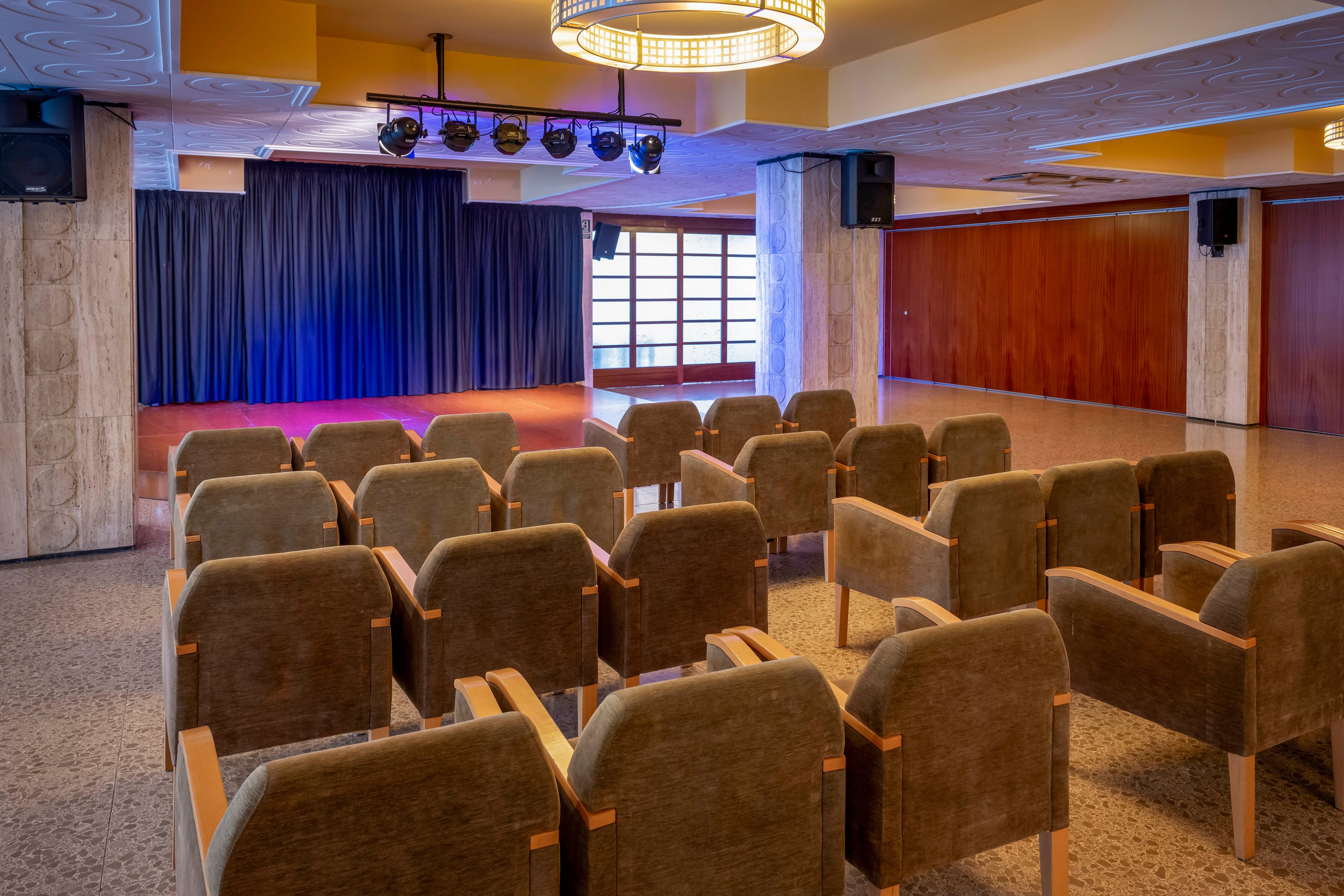 An empty conference room is arranged with rows of upholstered chairs facing a small stage. The stage features a dark curtain backdrop and overhead spotlights casting a blue and purple glow. Warm wooden wall panels and stone columns frame the space, giving it a modern yet cozy atmosphere. Large windows at the back allow natural light to complement the interior lighting.