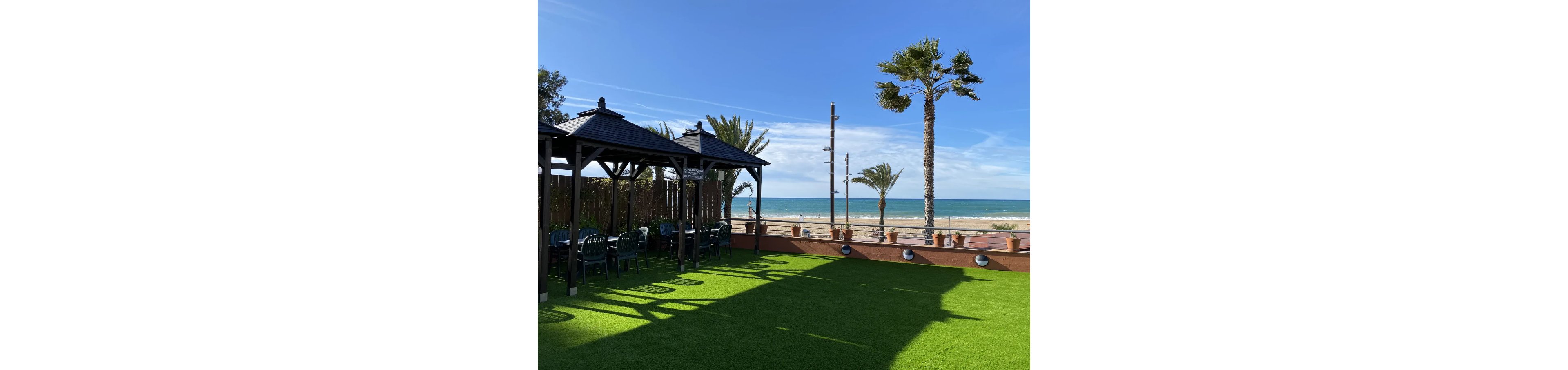 An outdoor terrace with wooden pergolas and plastic chairs overlooks a sandy beach and calm sea. The foreground features bright green artificial grass under a clear blue sky. Palm trees line the edge of the terrace, adding a tropical feel to the coastal setting. The scene suggests a relaxed, sunny environment ideal for leisure or dining by the ocean.