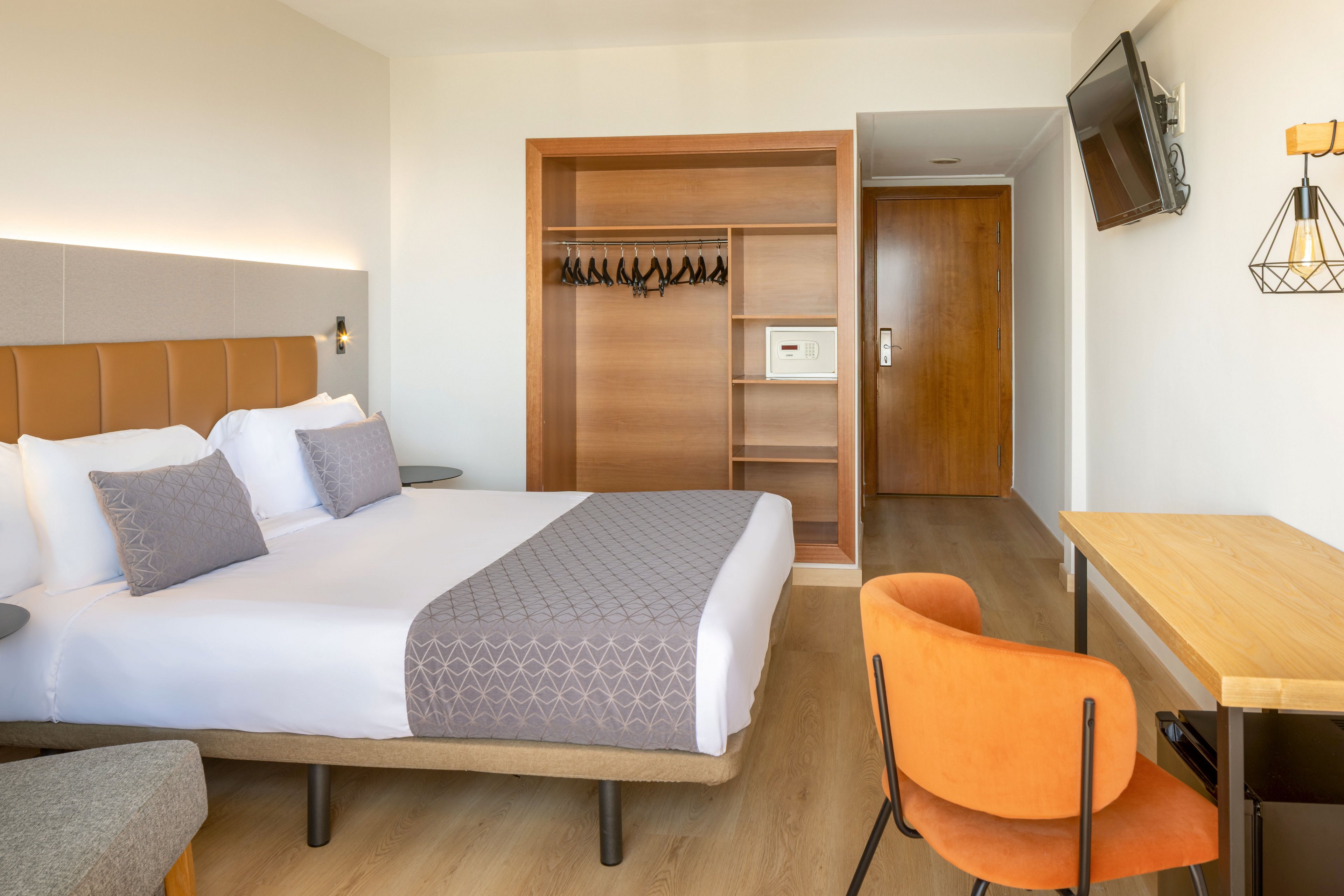 Contemporary hotel room featuring a neatly made double bed with white linens and patterned gray accents. The space includes an open wooden wardrobe with hangers and a wall-mounted safe near the entrance door. A wooden desk with an orange upholstered chair faces a wall-mounted television, adding functionality for work or leisure. Light-colored flooring and neutral tones create a bright, clean, and inviting atmosphere.