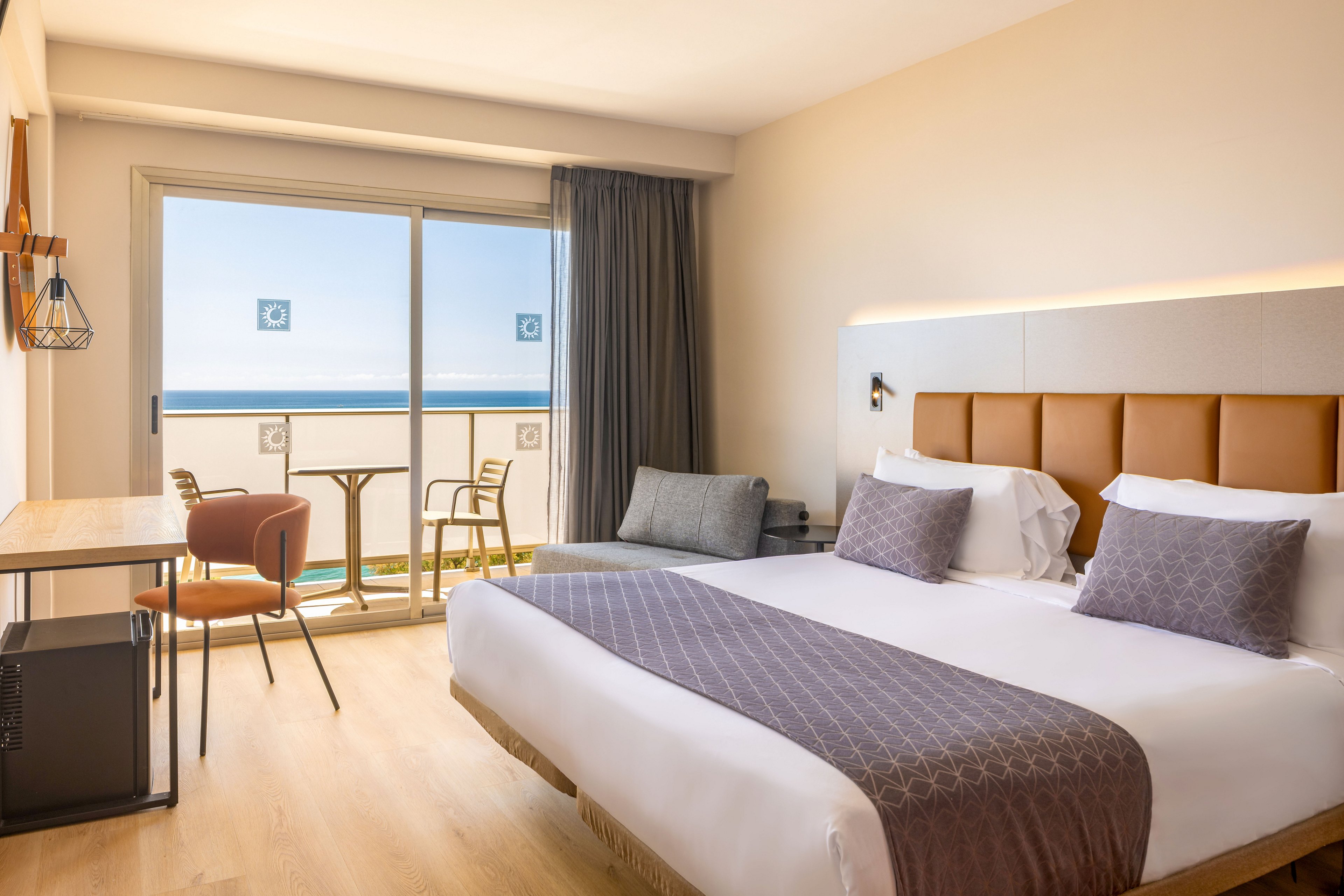 Contemporary hotel room featuring a large double bed with white linens and patterned runner. A private balcony with two chairs and a small table overlooks a clear blue ocean and sky. The interior includes a wooden desk with chair, a compact fridge, and a gray sofa, all in a neutral, minimalist style. Natural light floods the space through the sliding glass doors, creating a calm and inviting atmosphere.