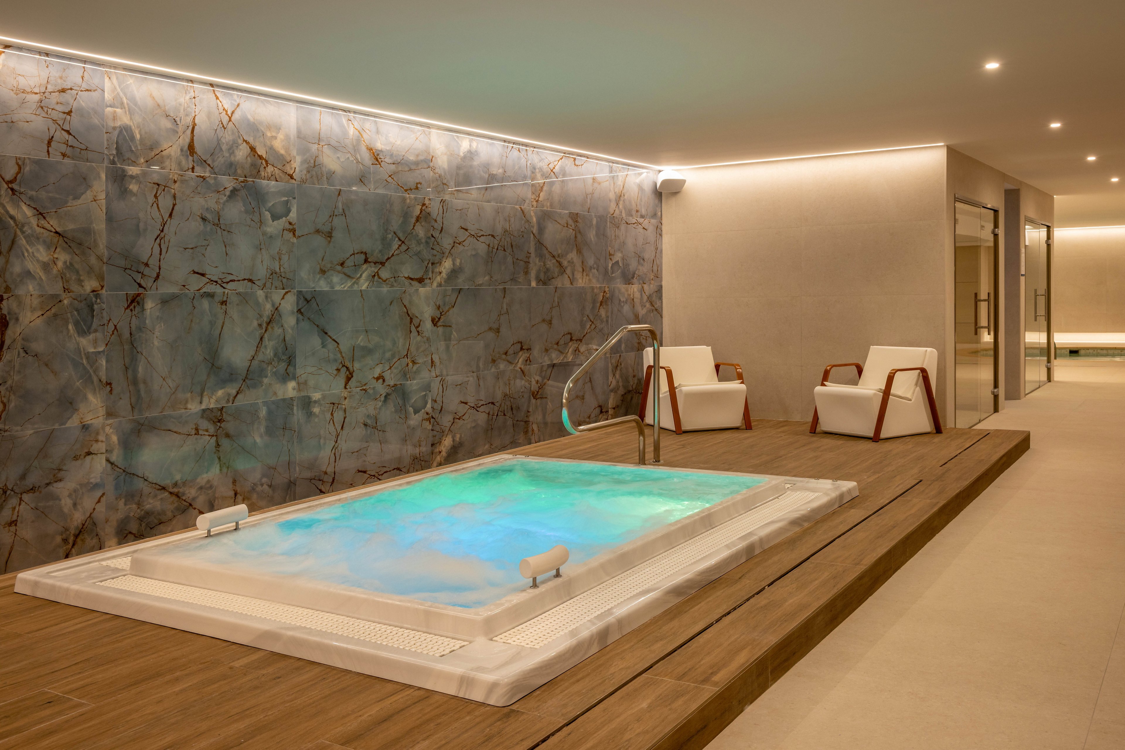 A contemporary indoor spa features a built-in jacuzzi with softly glowing turquoise water. The hot tub is set into a wooden platform, contrasting with the smooth light-colored flooring. Two modern white lounge chairs with wooden armrests sit nearby for relaxation. A large stone-textured wall and subtle recessed lighting create a calm, luxurious atmosphere.