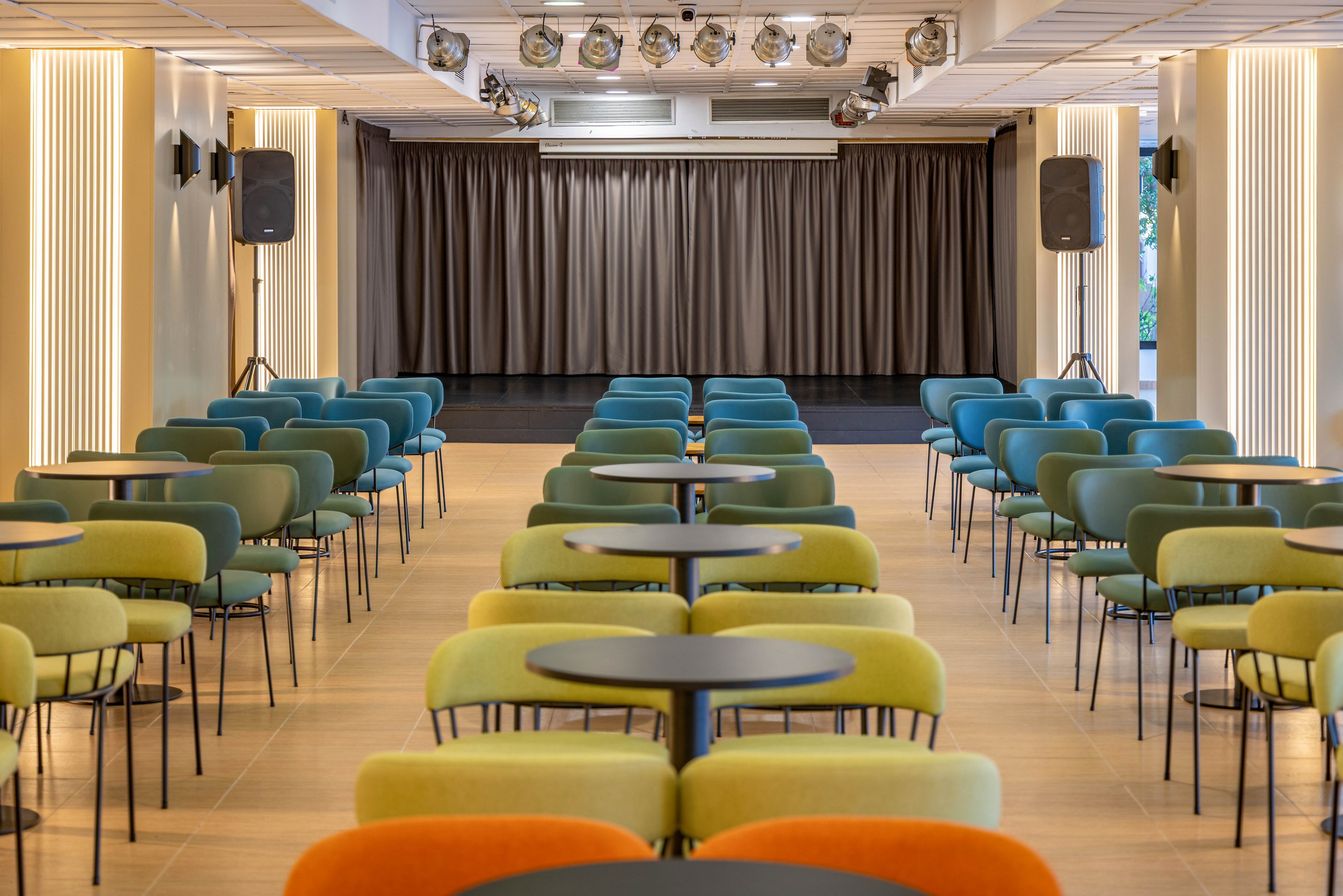 A contemporary conference room is arranged with rows of colorful chairs and round tables facing a stage. The space features a neutral floor, vertical light panels, and large speakers on either side of the stage. Overhead spotlights illuminate the area, highlighting the clean and organized layout. The room appears ready for a presentation, seminar, or corporate event.