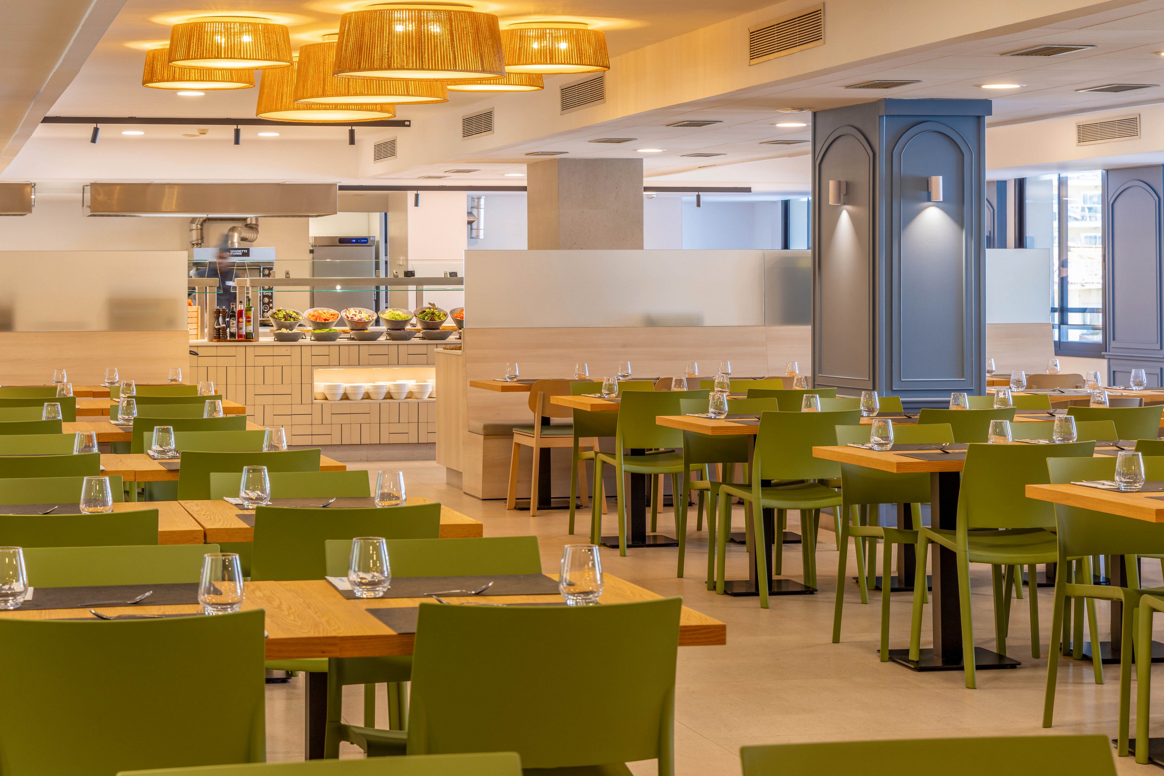A contemporary restaurant dining area features rows of wooden tables paired with green chairs. Each table is neatly set with placemats and upside-down water glasses, ready for guests. Warm pendant lights with woven shades hang from the ceiling, creating a cozy atmosphere. In the background, a buffet or serving counter displays fresh food under bright lighting, enhancing the clean and inviting interior design.