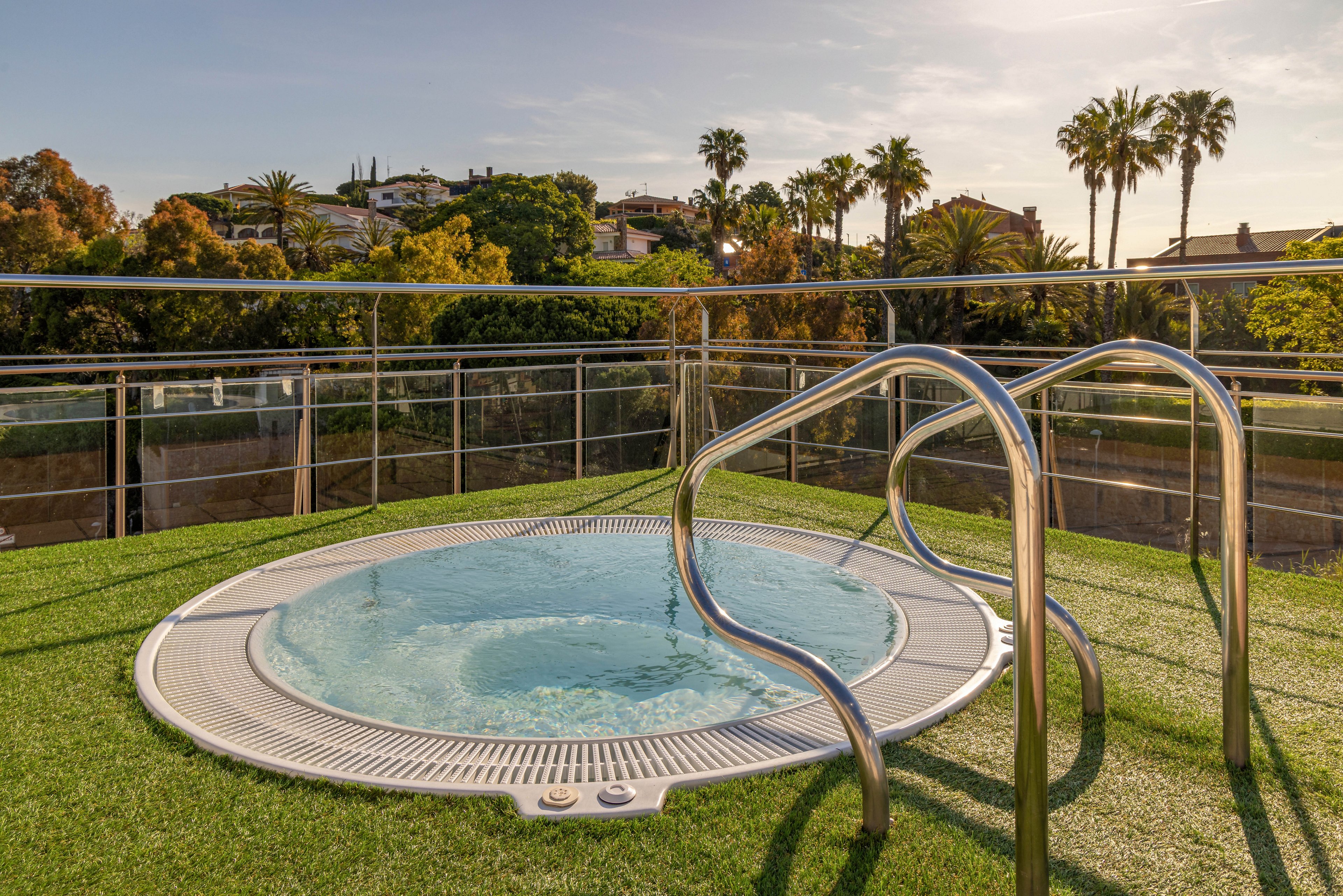 An outdoor circular jacuzzi sits on a rooftop terrace covered with artificial grass. Stainless steel handrails curve into the bubbling water, framed by a safety railing around the deck. In the background, palm trees and houses are visible under a warm, late afternoon sky. The scene conveys a relaxed, resort-like atmosphere ideal for leisure and wellness.