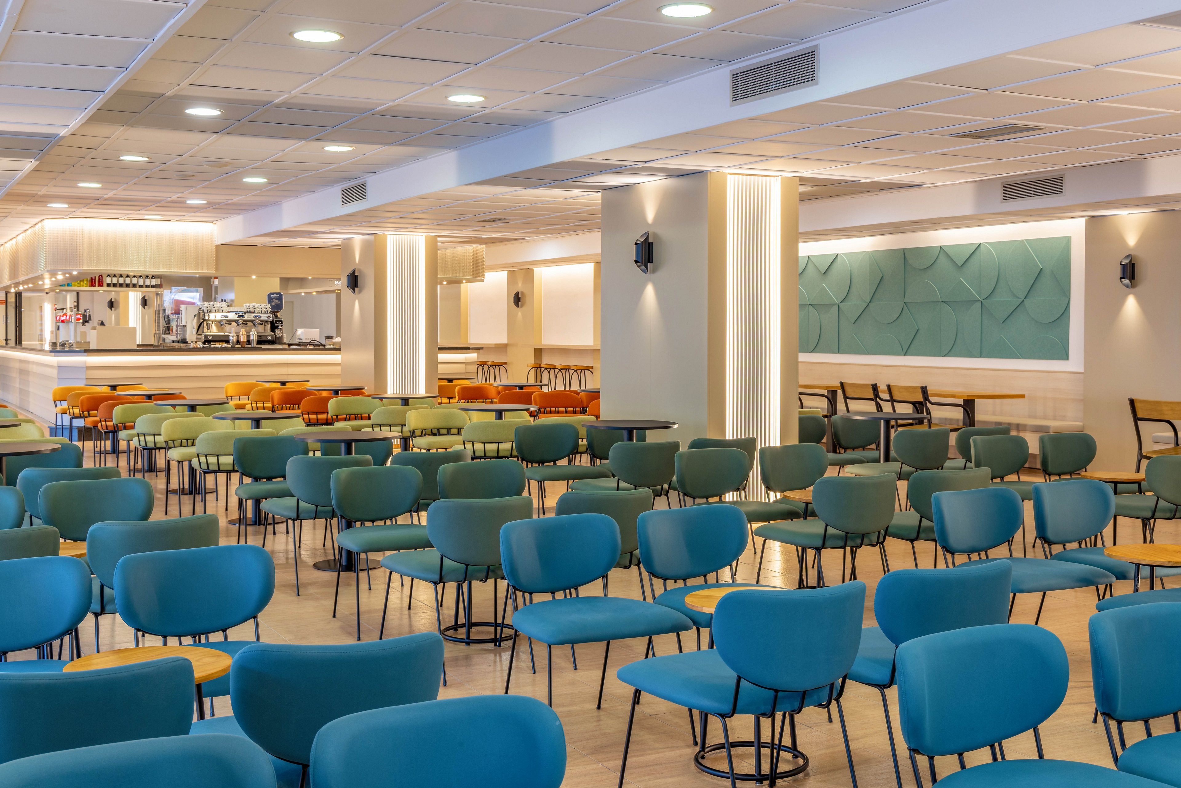 A spacious, modern lounge area is filled with rows of colorful chairs arranged in front of a bar. The setting appears to be a conference or event space, currently empty and ready for guests. Soft, warm lighting and light wood flooring create a welcoming atmosphere. Teal wall art and upholstered seating add a contemporary touch to the interior design.