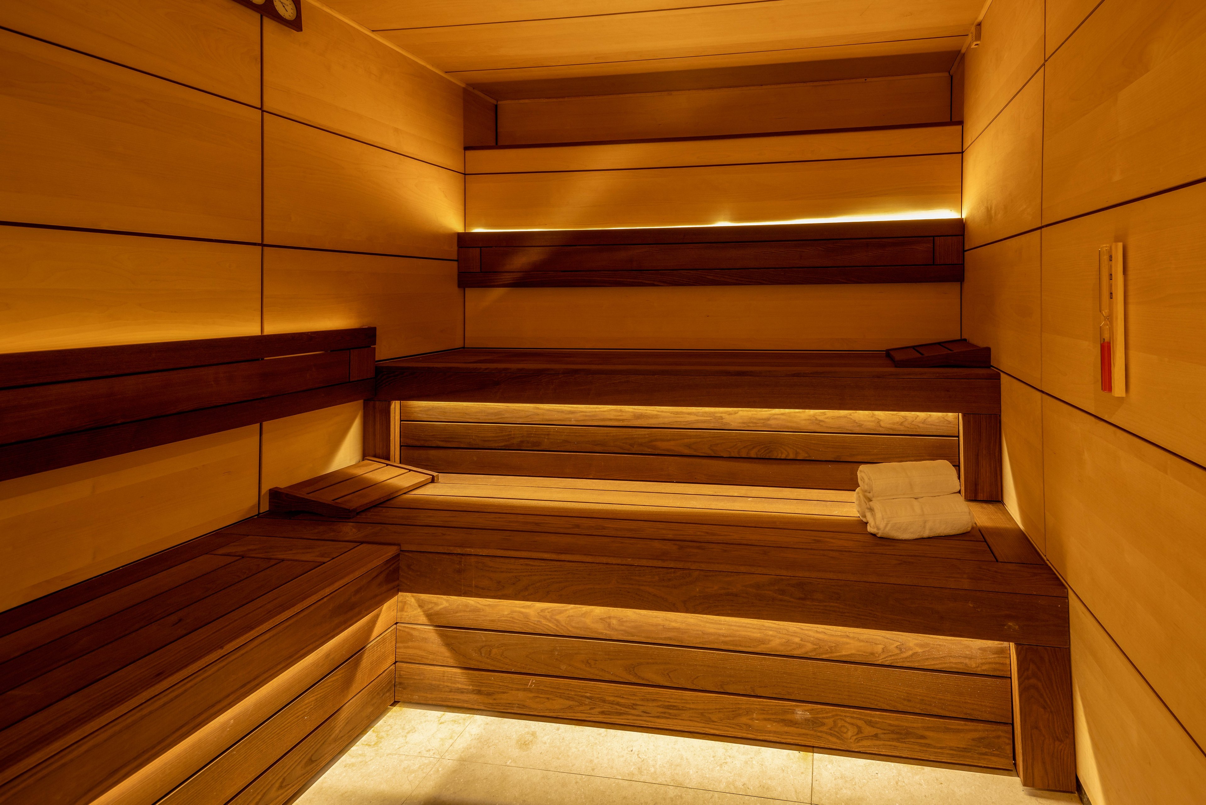 A modern wooden sauna interior is shown with tiered benches and soft indirect lighting. The room is empty, emphasizing clean lines and smooth wooden paneling on the walls and seating. A neatly folded white towel rests on one of the lower benches, suggesting readiness for use. The overall atmosphere feels warm, calm, and inviting, ideal for relaxation and wellness.