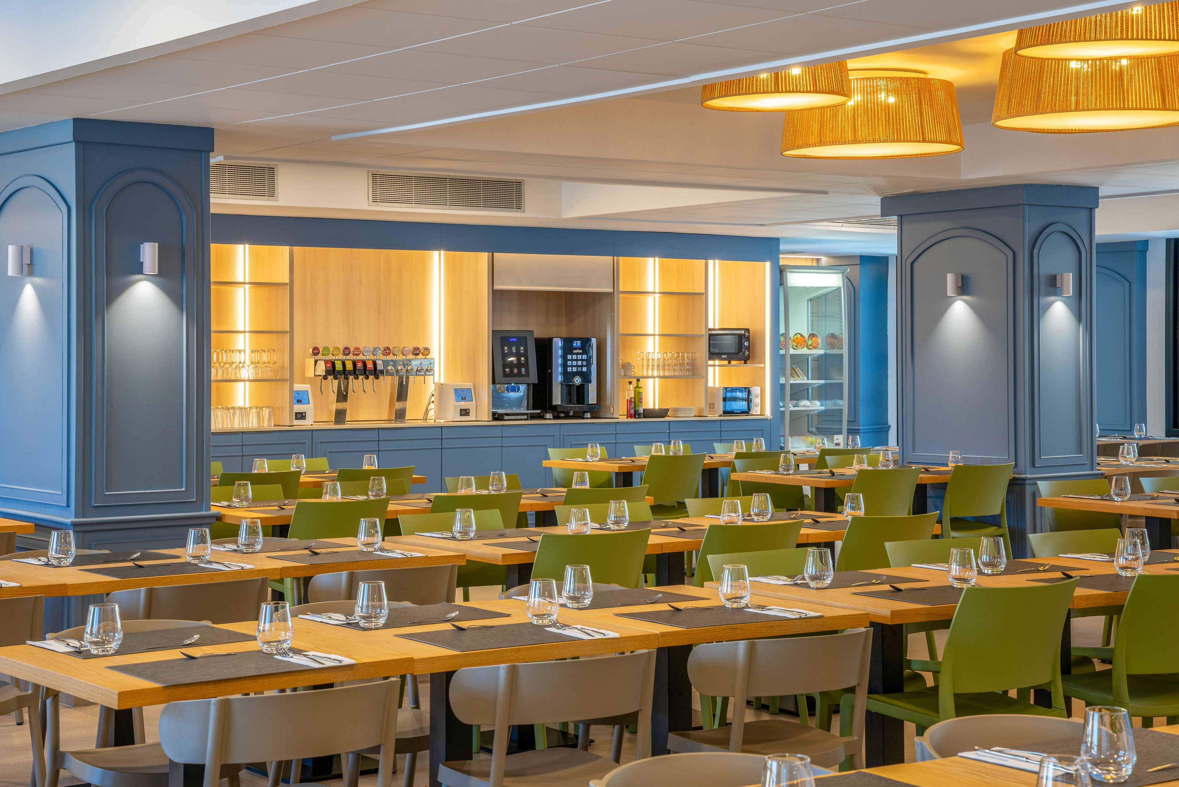 A contemporary restaurant dining area features long wooden tables neatly set with glasses and cutlery. Green and beige chairs are arranged in rows, creating an orderly and spacious seating layout. In the background, a well-lit service counter with beverage machines and shelves adds a functional focal point. Warm pendant lights and soft wall sconces contribute to a welcoming, modern atmosphere.