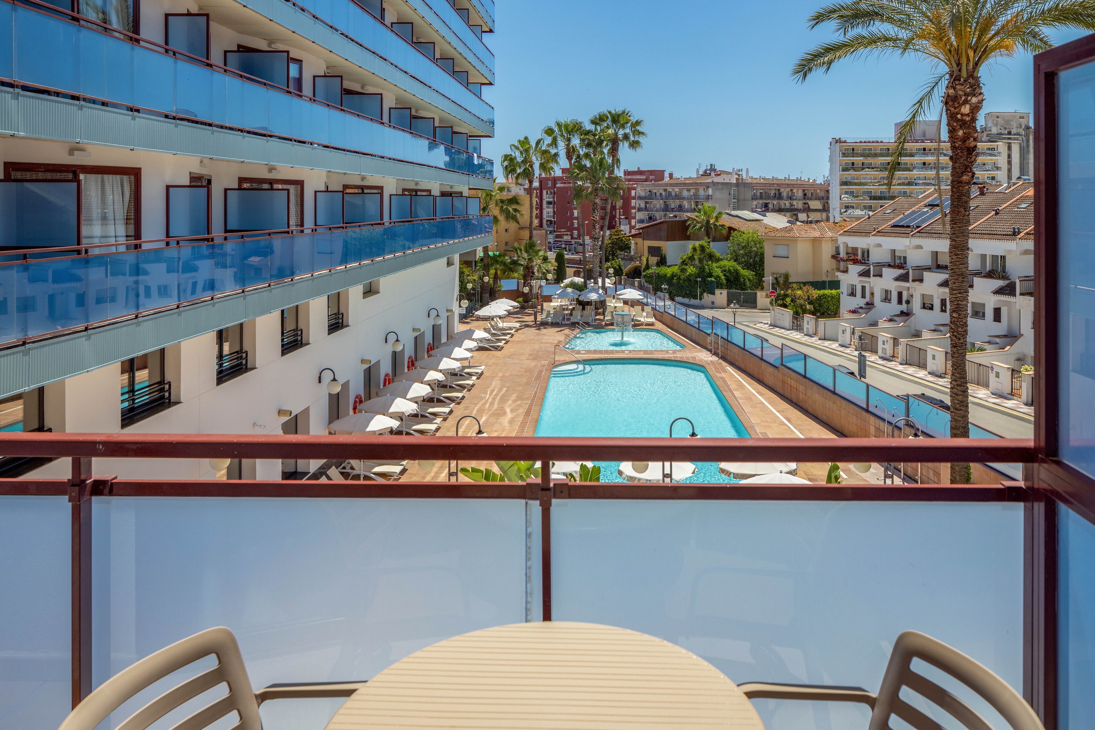 View from a hotel balcony looks out over a long outdoor swimming pool. Sun loungers and umbrellas line the poolside area between two modern apartment-style buildings. A small round table and chairs sit in the foreground on the balcony. Palm trees, clear blue sky, and distant city buildings create a bright, summery vacation atmosphere.