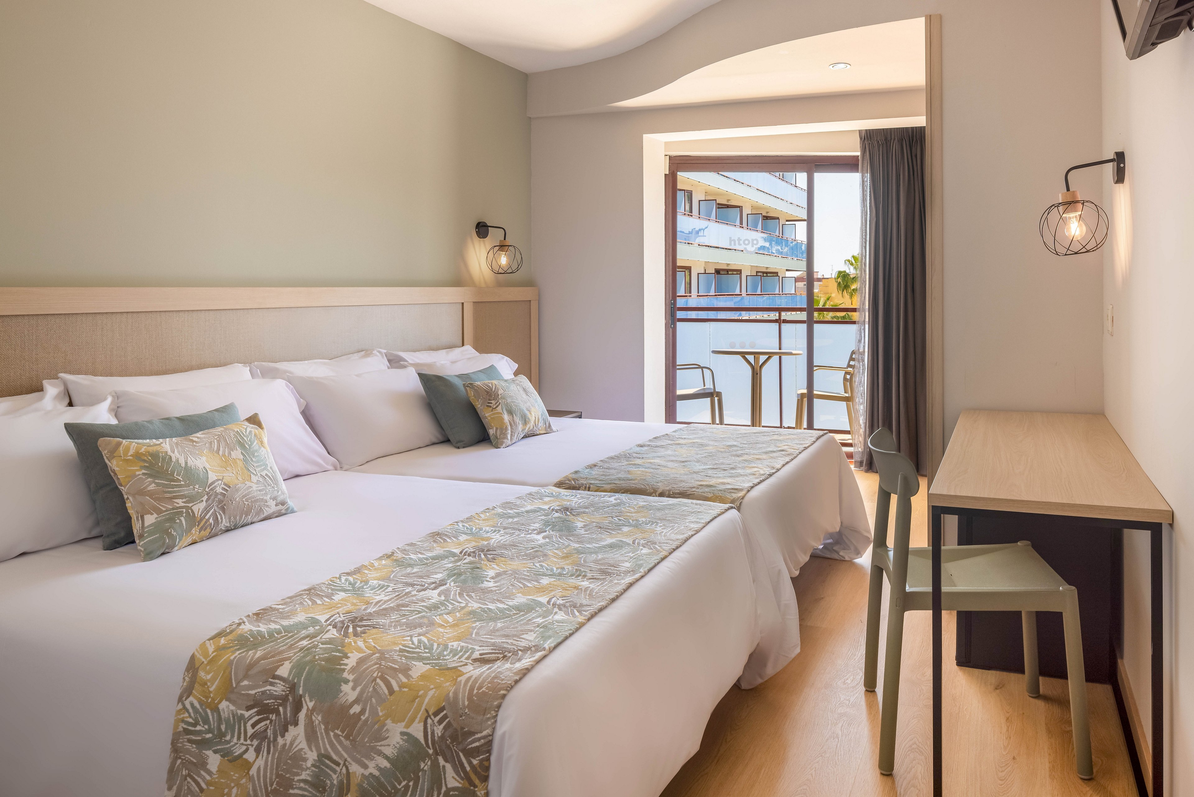 A modern hotel room features two neatly made beds with patterned runners and matching cushions. Soft neutral walls and wooden flooring create a calm, contemporary atmosphere. A small desk and chair sit beside the beds, while a glass door opens onto a balcony with outdoor seating. Through the balcony, another building and a glimpse of water or pool area are visible, suggesting a resort setting.