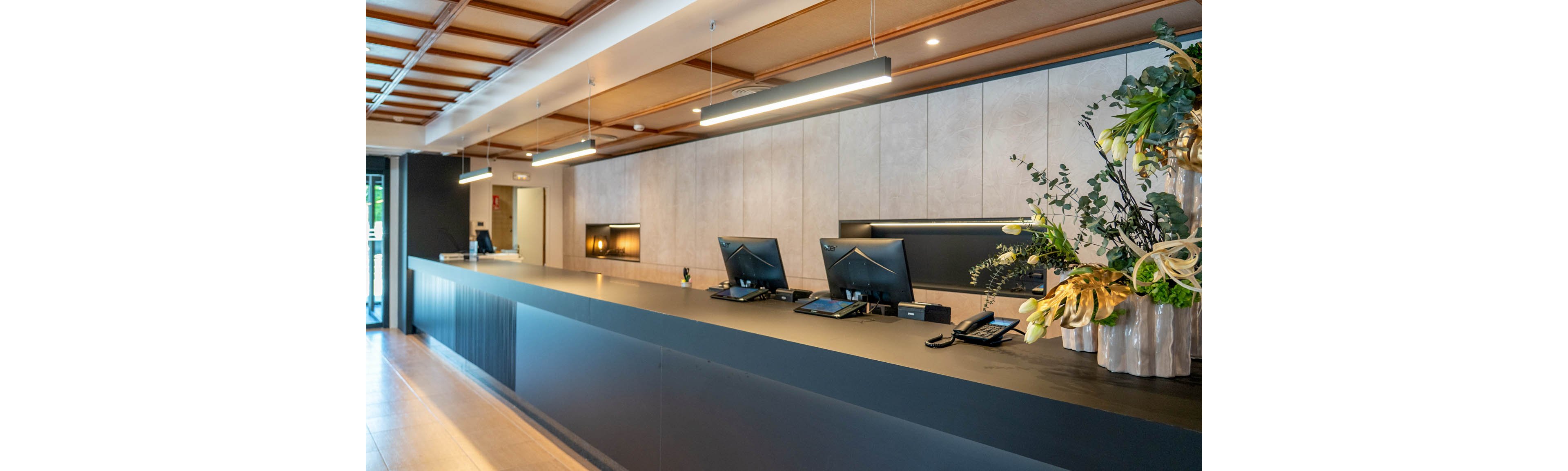 A long, modern reception counter stretches across a bright lobby interior. Two computer workstations and telephones sit neatly on the dark countertop, suggesting a hotel or office front desk. Warm ceiling lights and wood paneling create a welcoming atmosphere. A large floral arrangement on the right adds a decorative touch to the clean, minimalist space.