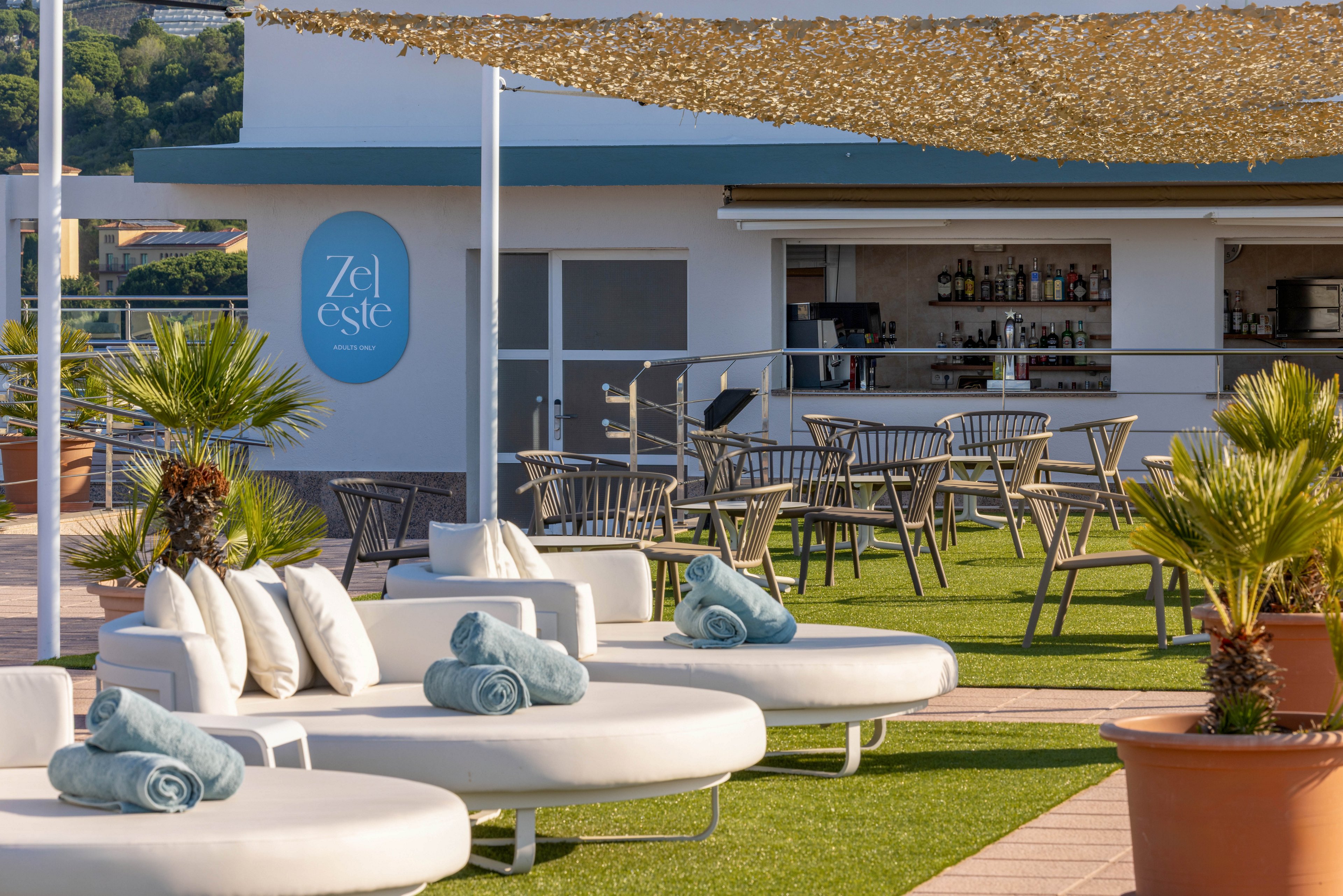 A stylish rooftop lounge features round white sunbeds with rolled blue towels and cushions arranged on artificial grass. In the background, an open-air bar with wooden tables and chairs sits beneath a shade canopy. Large potted plants and small palm trees add a tropical touch to the modern setting. A circular wall sign reading "Zel Este" is visible on the bar building.